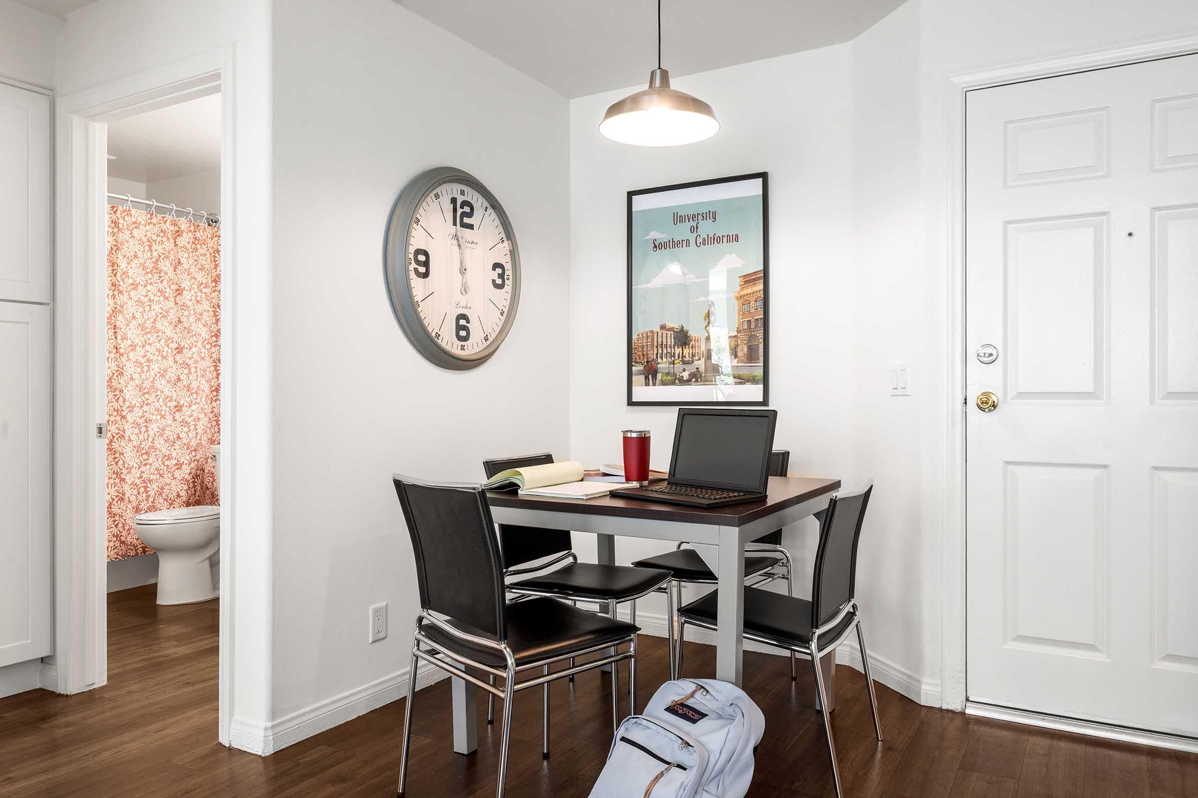 A small dining area featuring a round table with four black chairs, an open laptop, a notebook, and a drink. A large wall clock is shown, along with a framed poster of the University of Southern California. A doorway leads to a bathroom, and the floor has hardwood finishes.