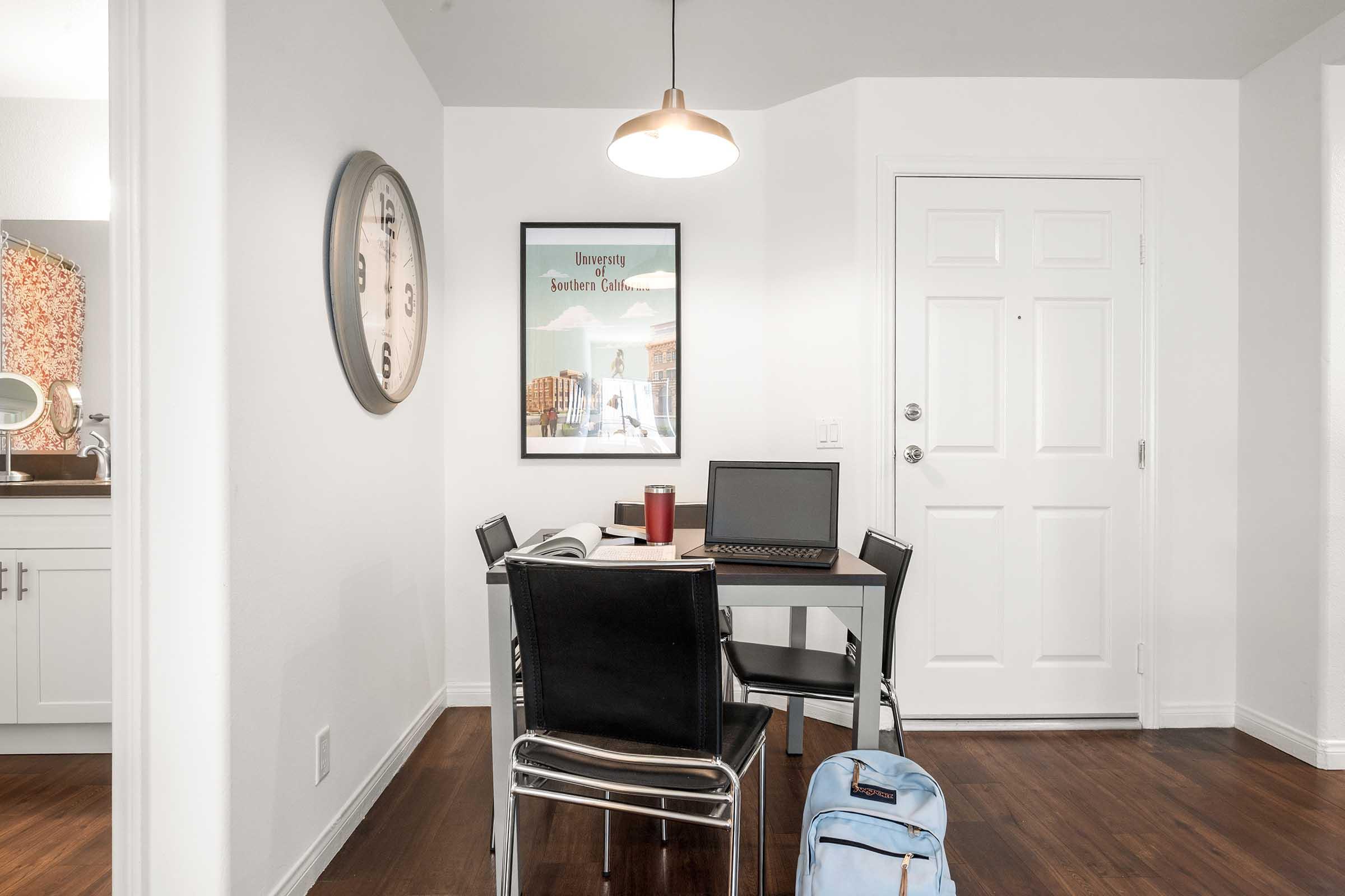 A bright, modern living space featuring a small dining table with a laptop, a red cup, and black chairs. A wall clock and an art piece are visible, along with a partially open door leading to another room. A light fixture hangs from the ceiling, and a blue backpack is positioned near the table.