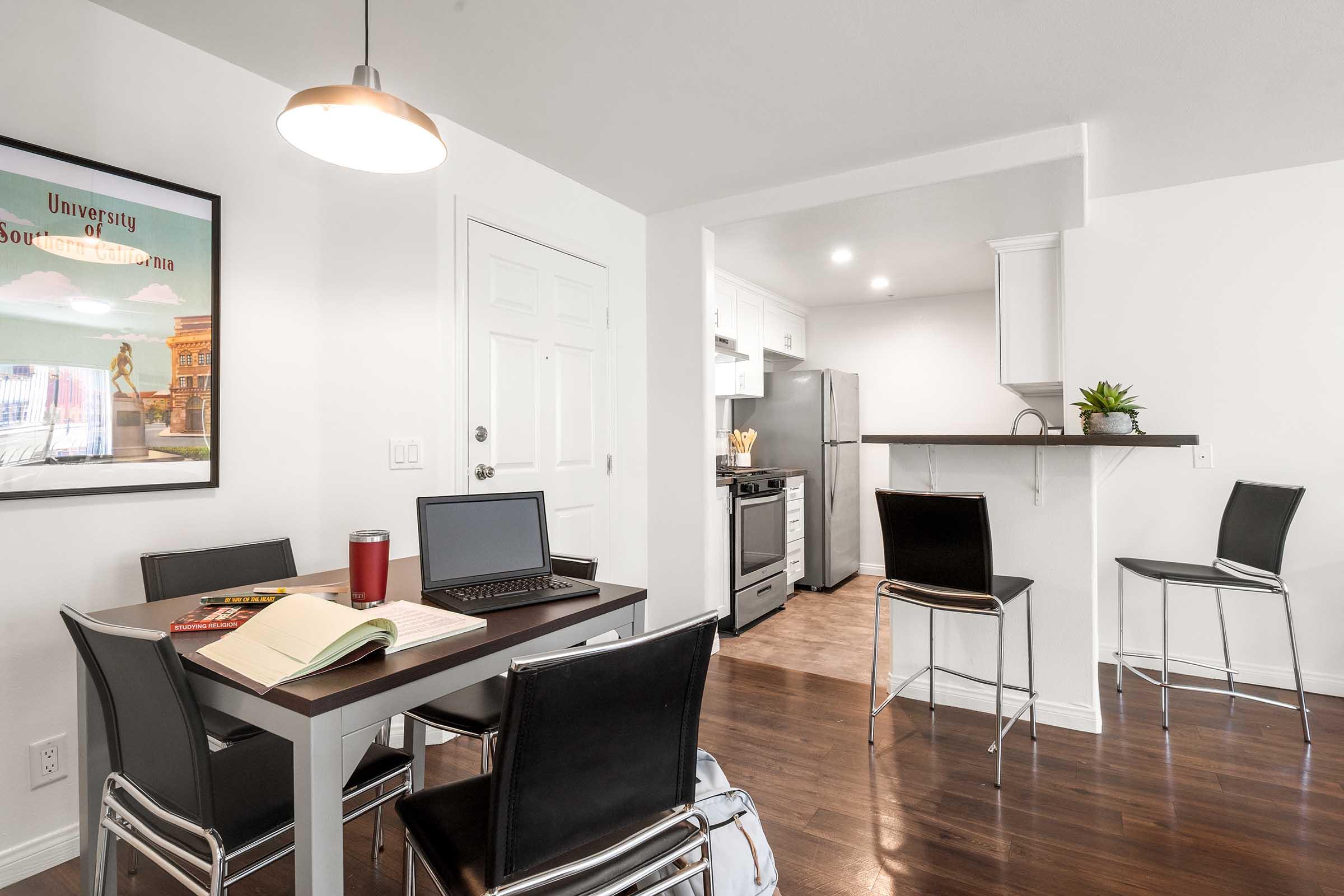 A modern kitchen and dining area featuring a small black table with a laptop, notebook, and a travel mug. There are four black chairs around the table. The kitchen includes a stove, refrigerator, and white cabinets, with a wall-mounted plant for decoration. A framed poster is visible on the wall.