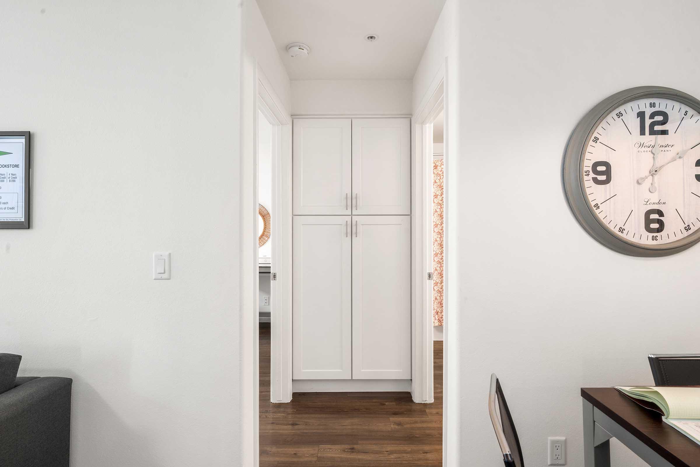 A view of a modern hallway featuring white cabinetry, leading to a room at the end. On the right, there's a dining table with a chair. A round wall clock is mounted on the wall, indicating the time. The flooring is dark wood, and the walls are painted white, creating a clean and bright atmosphere.