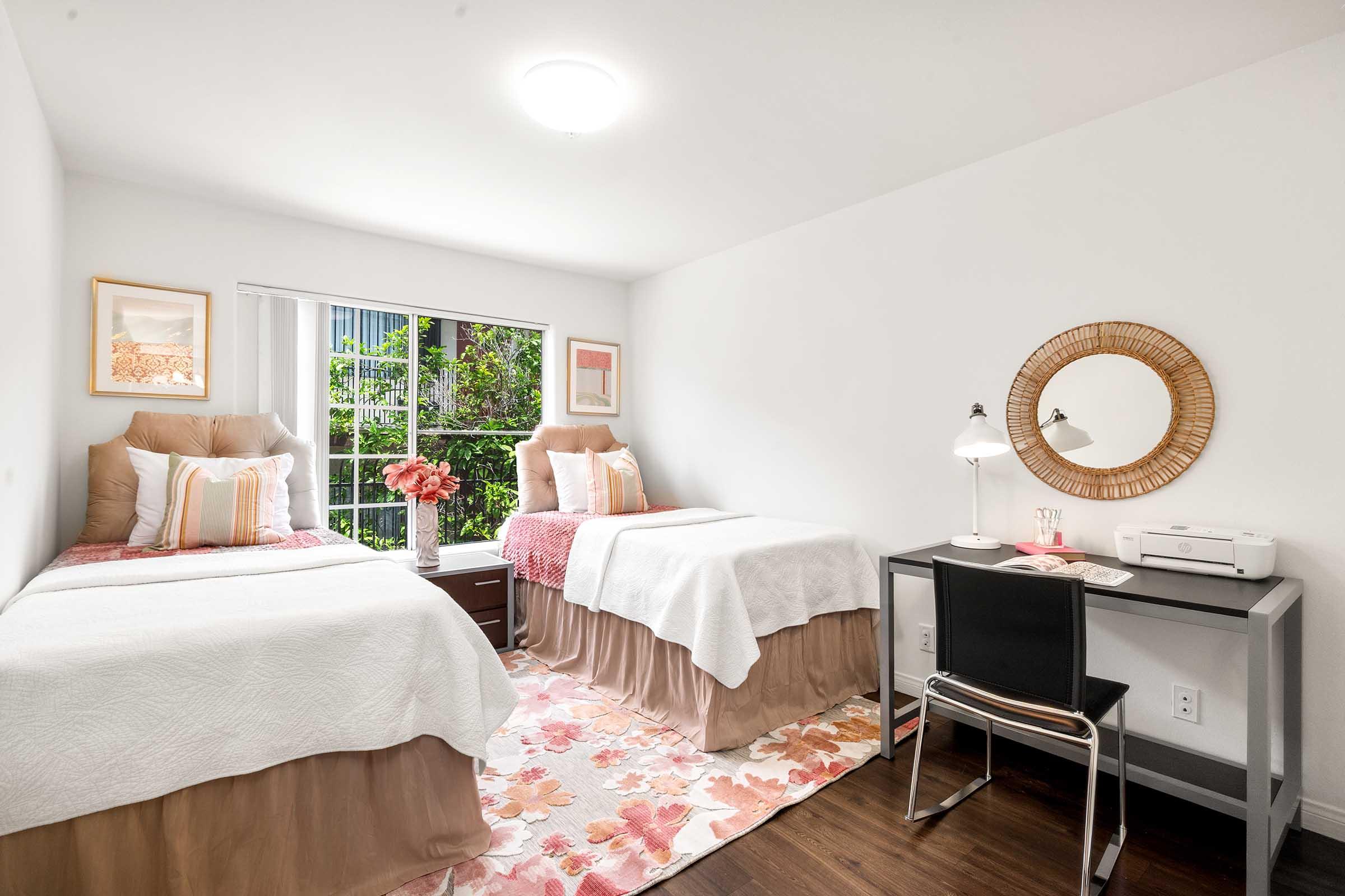 A brightly lit room featuring two twin beds with decorative pillows, a desk with a lamp and printer, and a circular mirror on the wall. The decor includes a floral area rug and artwork, with greenery visible through the window. The overall color scheme is soft and inviting.