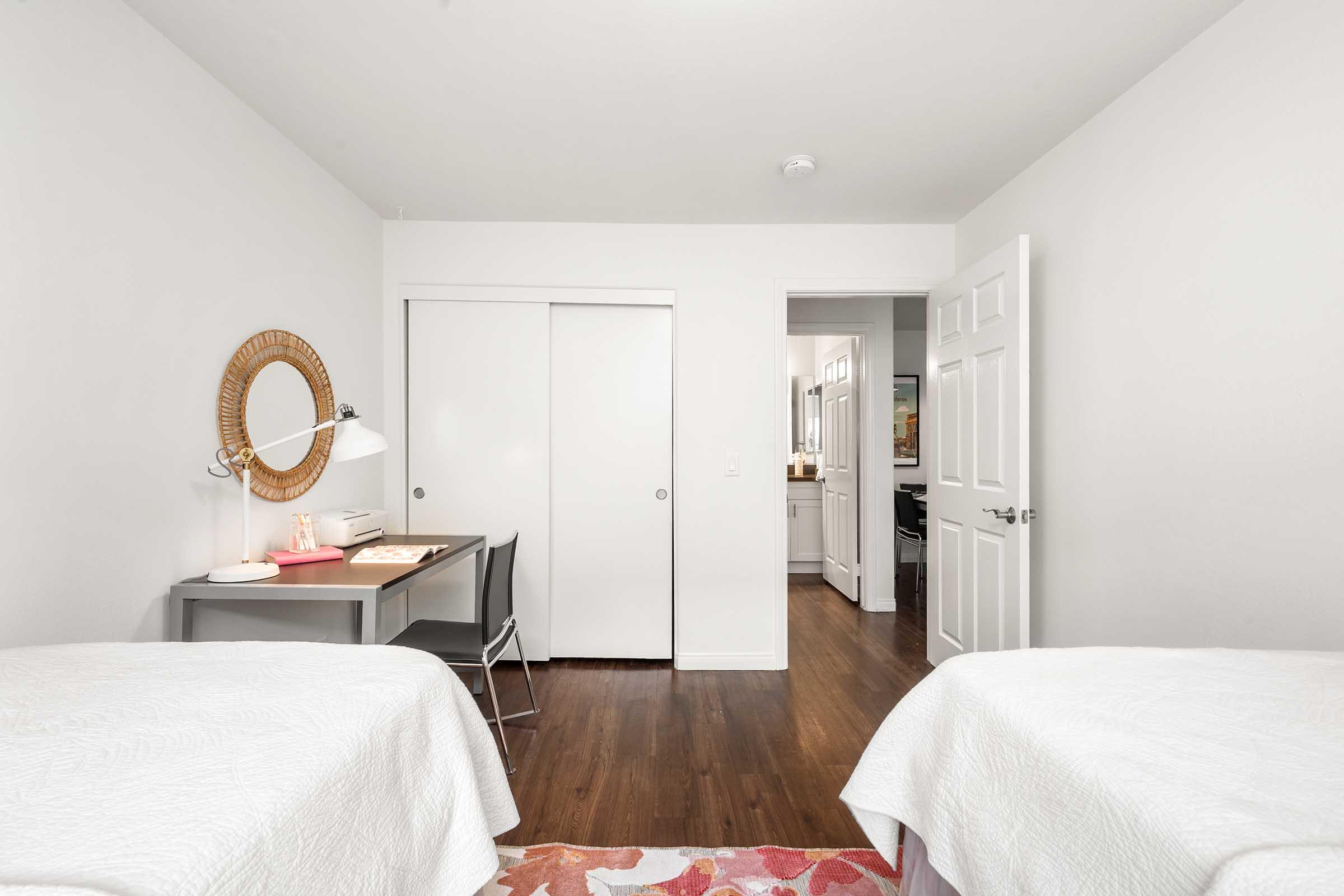A bright and modern bedroom featuring two twin beds with white bedspreads, a small desk with a lamp and stationery, a round mirror hanging on the wall, and a colorful area rug. A closet with sliding doors is visible, along with an open door leading to a bathroom or kitchen area.