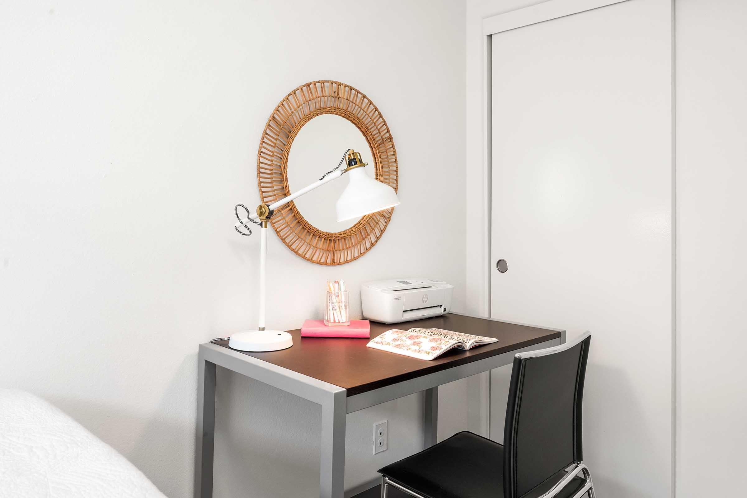 A minimalist workspace featuring a black desk with a white lamp, a modern mirror on the wall, a printer, and a magazine on the desk. A black chair is positioned at the desk, and a white closet door is visible in the background. The overall decor is clean and simple, emphasizing a bright atmosphere.