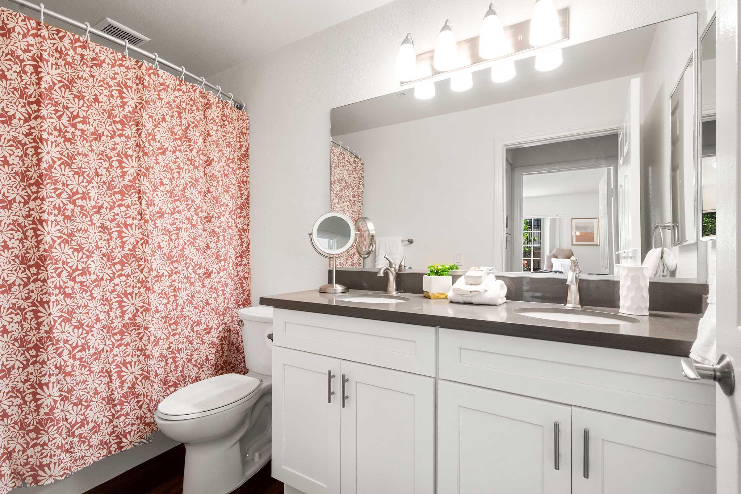 A modern bathroom featuring a white vanity with a dark countertop, a large mirror, and a decorative pink floral shower curtain. A toilet is visible, along with a small potted plant and a magnifying mirror on the counter. Soft lighting from above enhances the space's bright and clean aesthetic.