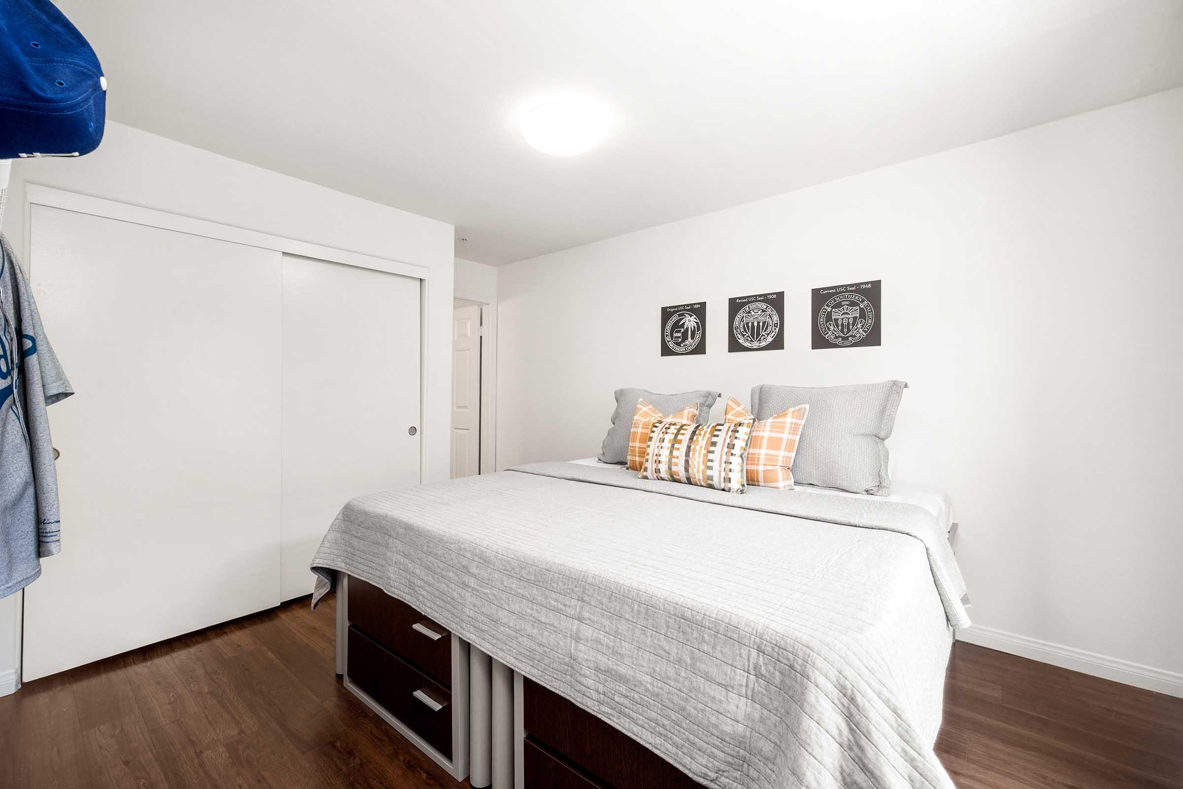 A cozy bedroom featuring a neatly made bed with gray bedding and decorative pillows. The walls are white, and above the bed are three framed artworks. There's a closet with sliding doors on one side and a blue cap hanging nearby, adding a personal touch to the space. The flooring is light wood.