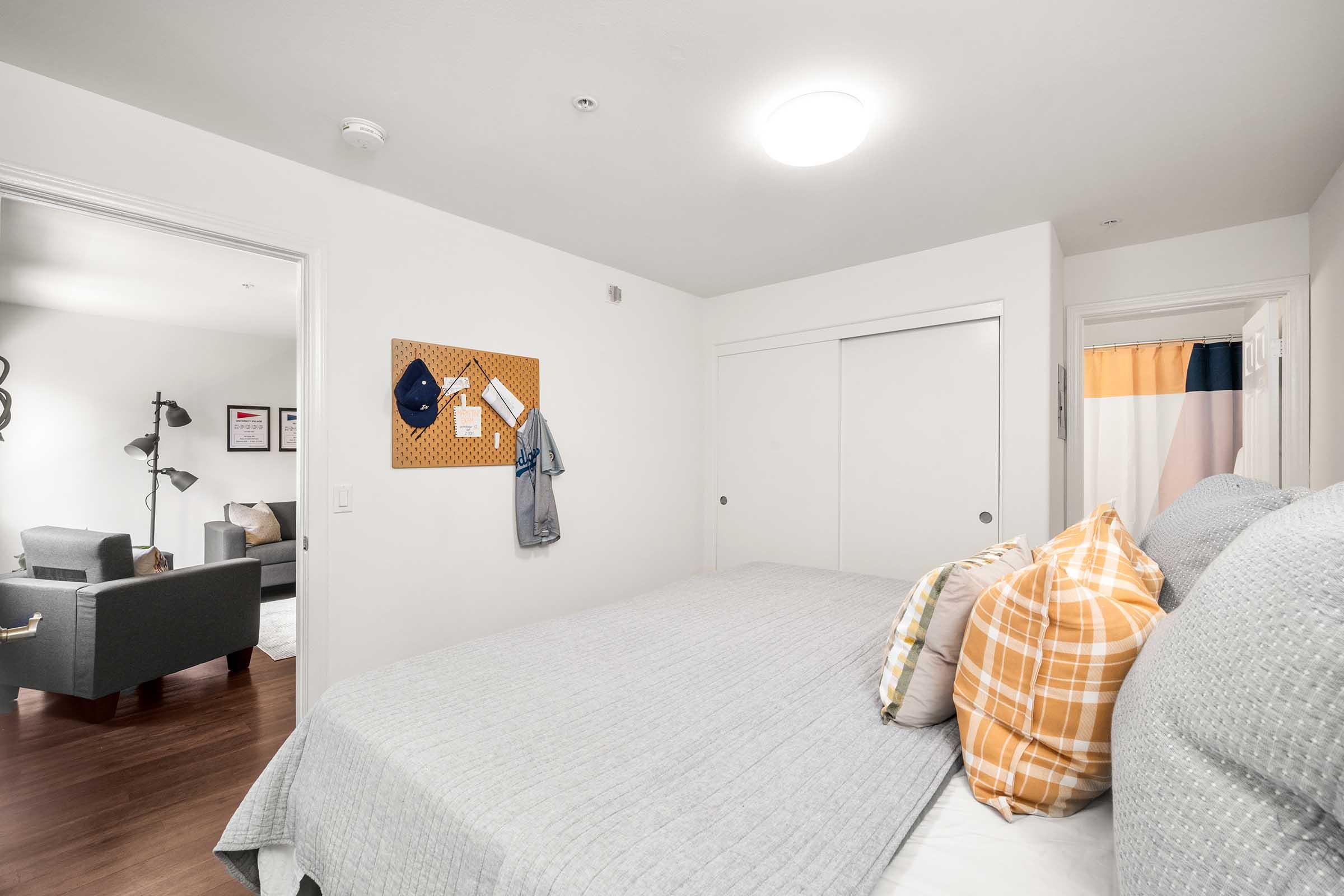 A cozy bedroom featuring a gray bedspread with decorative pillows in warm colors. A bulletin board on the wall holds a cap and clothing item. There's a closet with sliding doors, and visible through an open door is a modern living area with a gray armchair and light-colored rug. Natural light brightens the room.