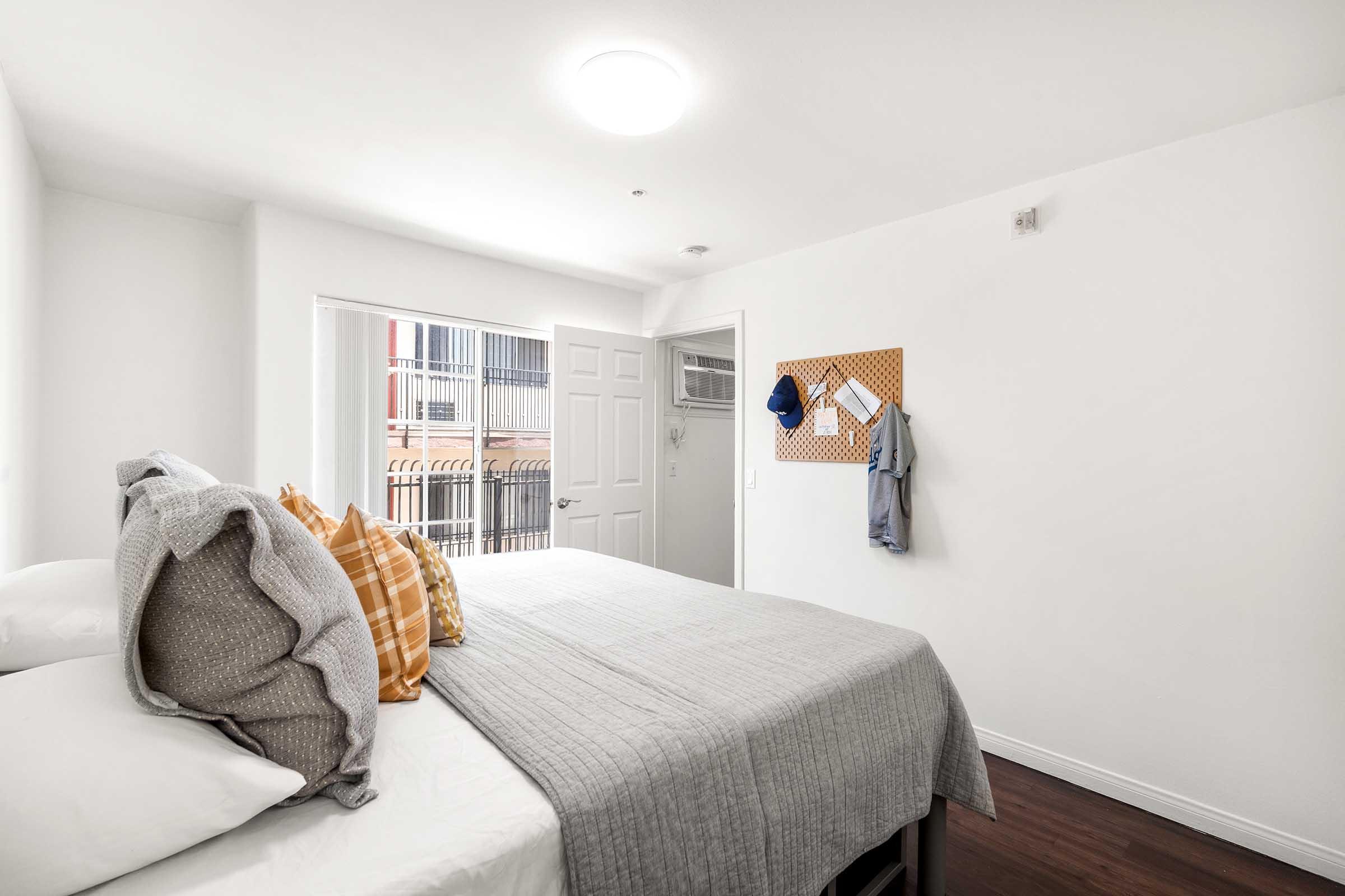 A bright and modern bedroom featuring a neatly made bed with decorative pillows, light-colored walls, and a wooden floor. A door leads to an outdoor area, and a pegboard on the wall displays a shirt and a hat. Natural light floods the room, creating a warm and inviting atmosphere.