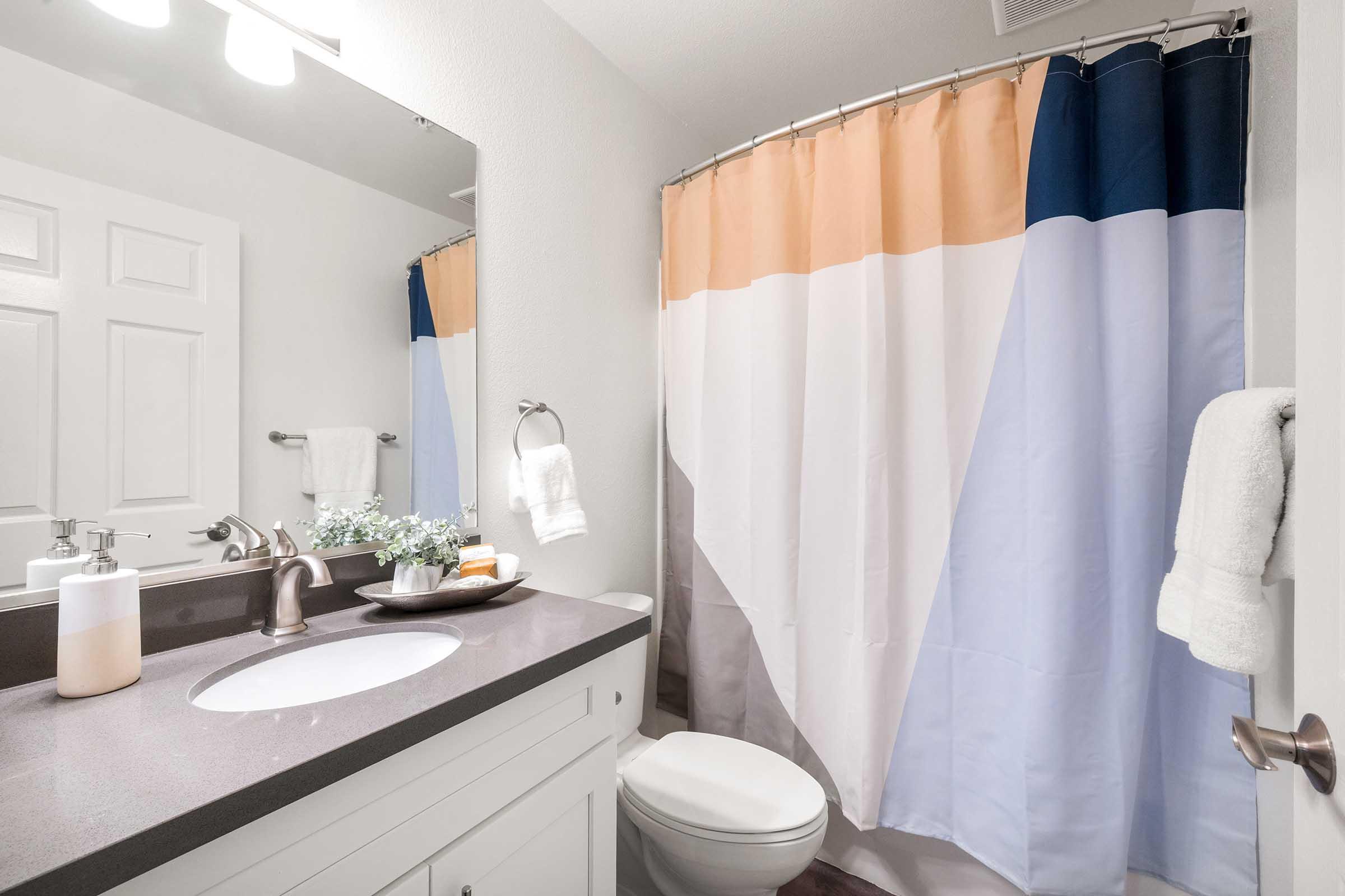 A modern bathroom featuring a clean design with a white vanity, a round sink, and a large mirror. The shower area is curtains with geometric patterns in muted colors. A stylish towel rack displays white towels, and a small plant and toiletries are arranged on the counter, adding a touch of warmth to the space.