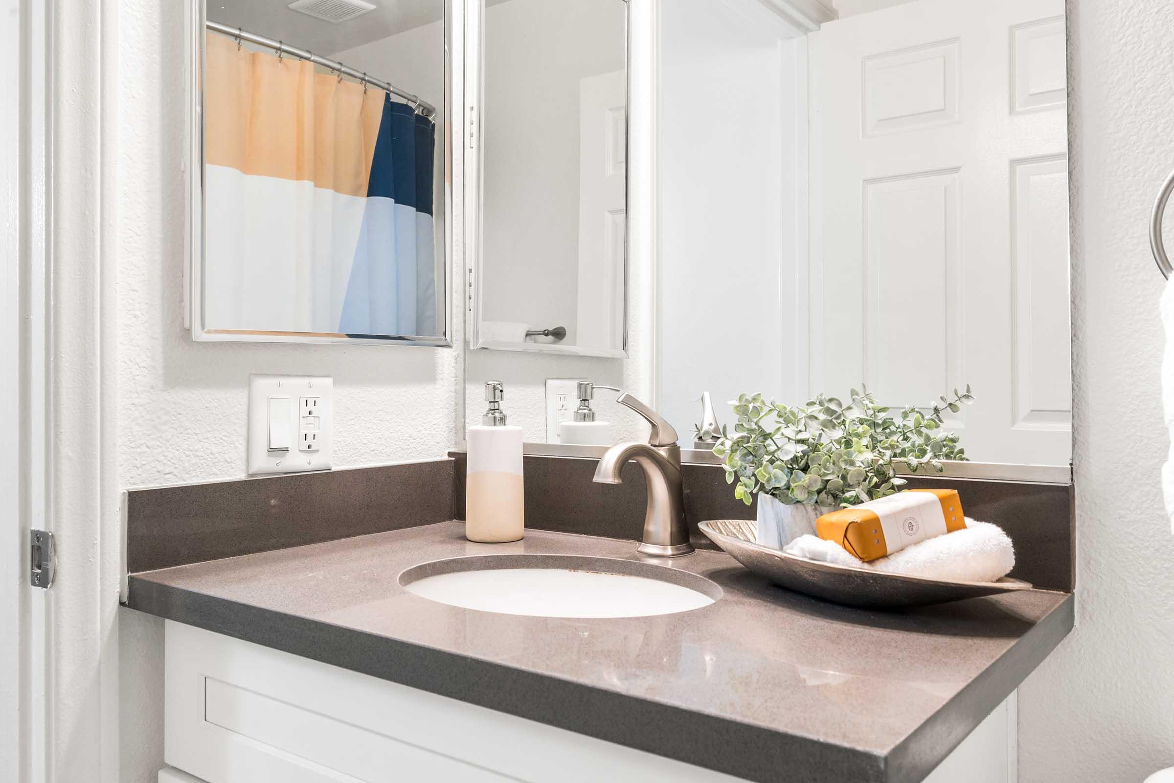 A modern bathroom vanity featuring a dark countertop with a sink, a soap dispenser, and a decorative dish holding a rolled towel and a small plant. Mirrors are mounted above the sink, and a colorful shower curtain is visible in the background. The space is well-lit and organized.