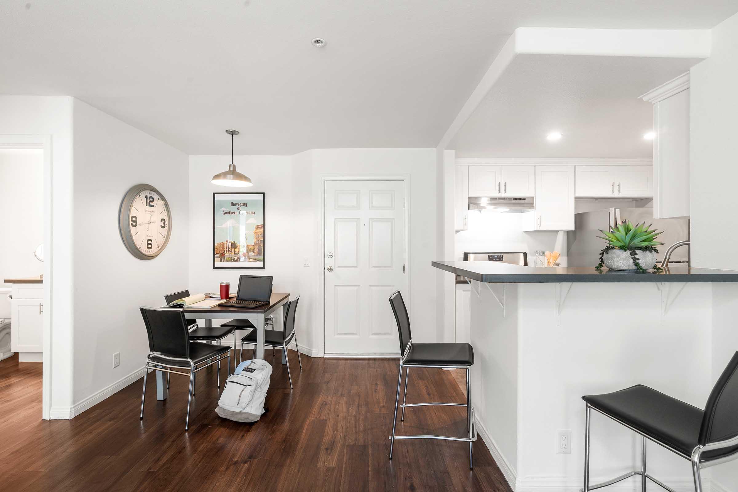 A well-lit, modern kitchen and dining area featuring a round table with chairs, a wall clock, and a backpack on the floor. The kitchen includes white cabinets, a countertop, and a door leading to another room. The space has hardwood flooring and a minimalist design.