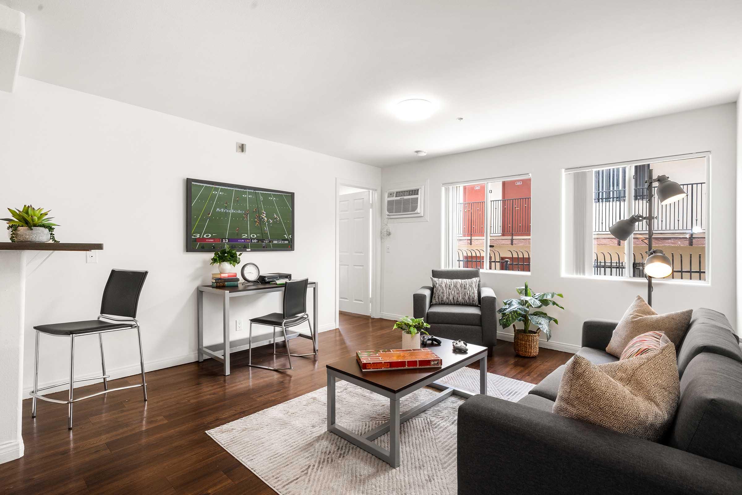 Modern living room with a dark sofa and armchairs, a coffee table with books and a plant, and a television mounted on the wall showing a football game. Two windows provide natural light, and an air conditioning unit is visible. The decor features neutral colors and a cozy, inviting atmosphere.