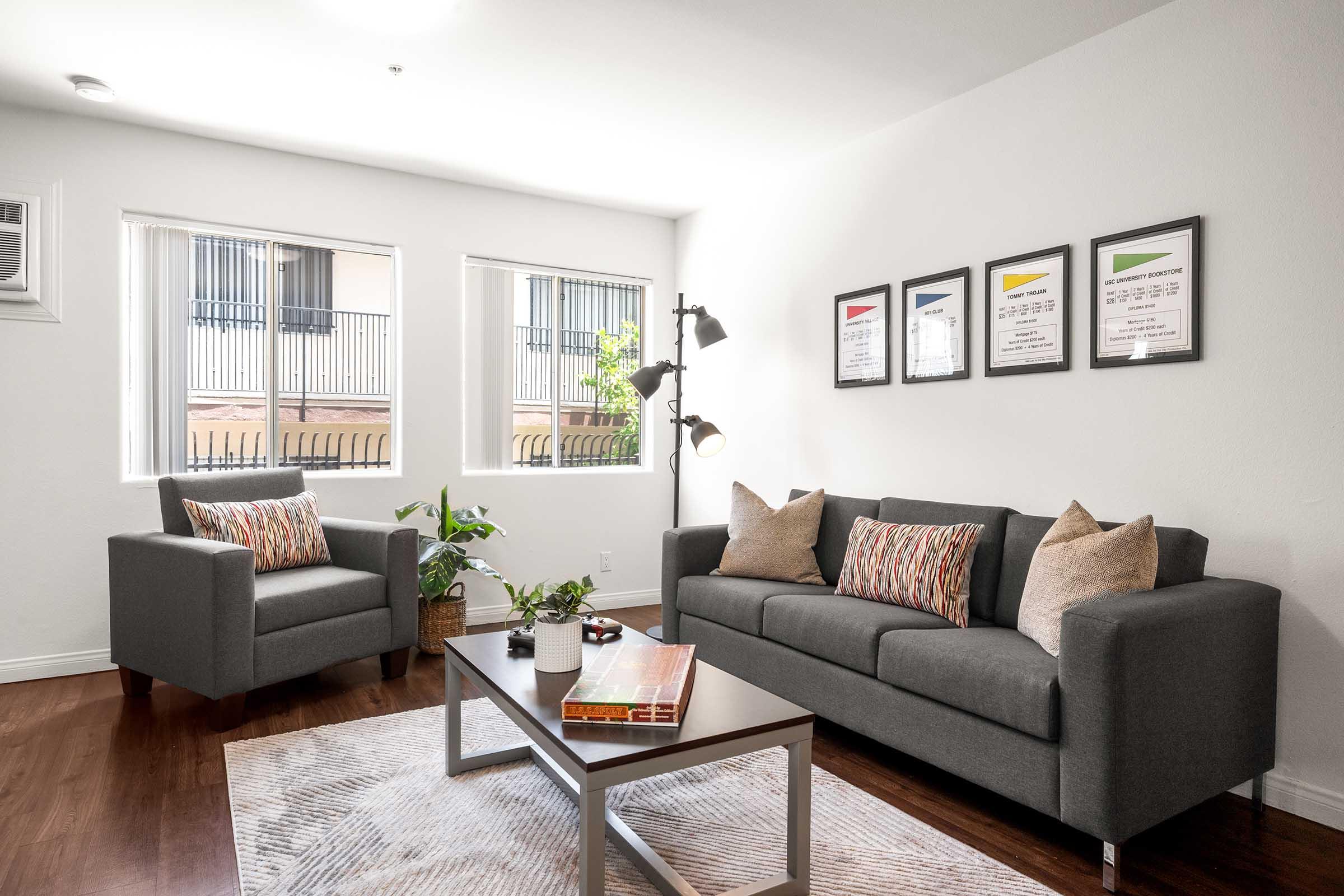 A modern living room featuring a gray sofa with decorative pillows, an armchair, and a wooden coffee table. The space has three framed art pieces on the wall, potted plants, and large windows allowing natural light. The flooring is hardwood, creating a cozy atmosphere.