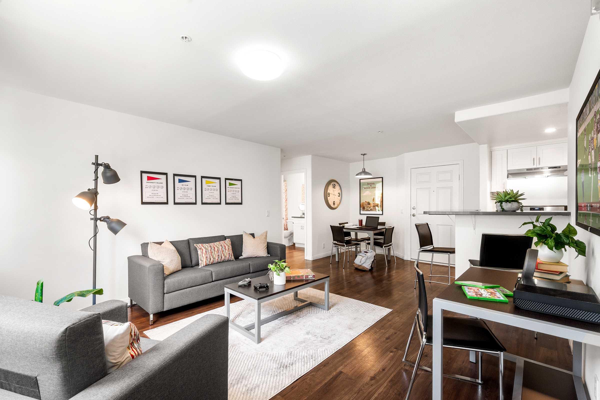 Modern living room with a gray sofa, decorative pillows, and a coffee table. Wall art featuring framed prints hangs above the sofa. A dining area with a small table and chairs is visible in the background, alongside a kitchen bar. The space has hardwood floors and bright lighting, creating a welcoming atmosphere.