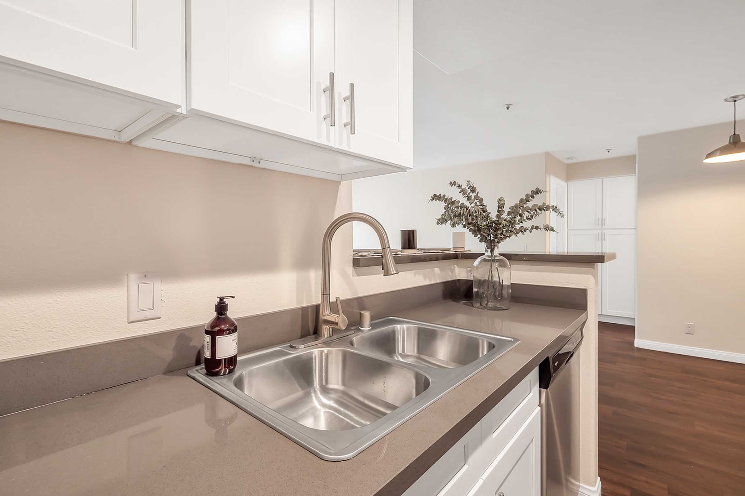 Modern kitchen with white cabinets, a double stainless steel sink, a sleek faucet, and a decorative vase with greenery on the countertop. The background features light-colored walls and wooden flooring.