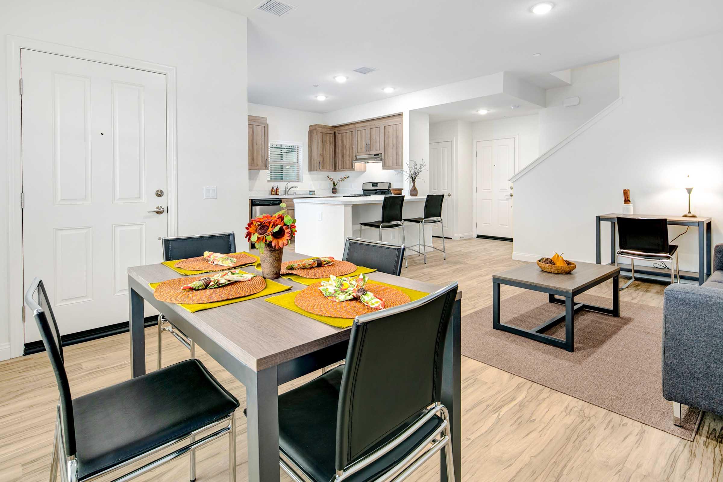 A modern open-plan living and dining area featuring a dining table set with colorful placemats and plates, a cozy living room with a sofa, and a kitchen in the background. The space has light-colored walls, wood cabinets, and warm flooring, creating a bright and inviting atmosphere.