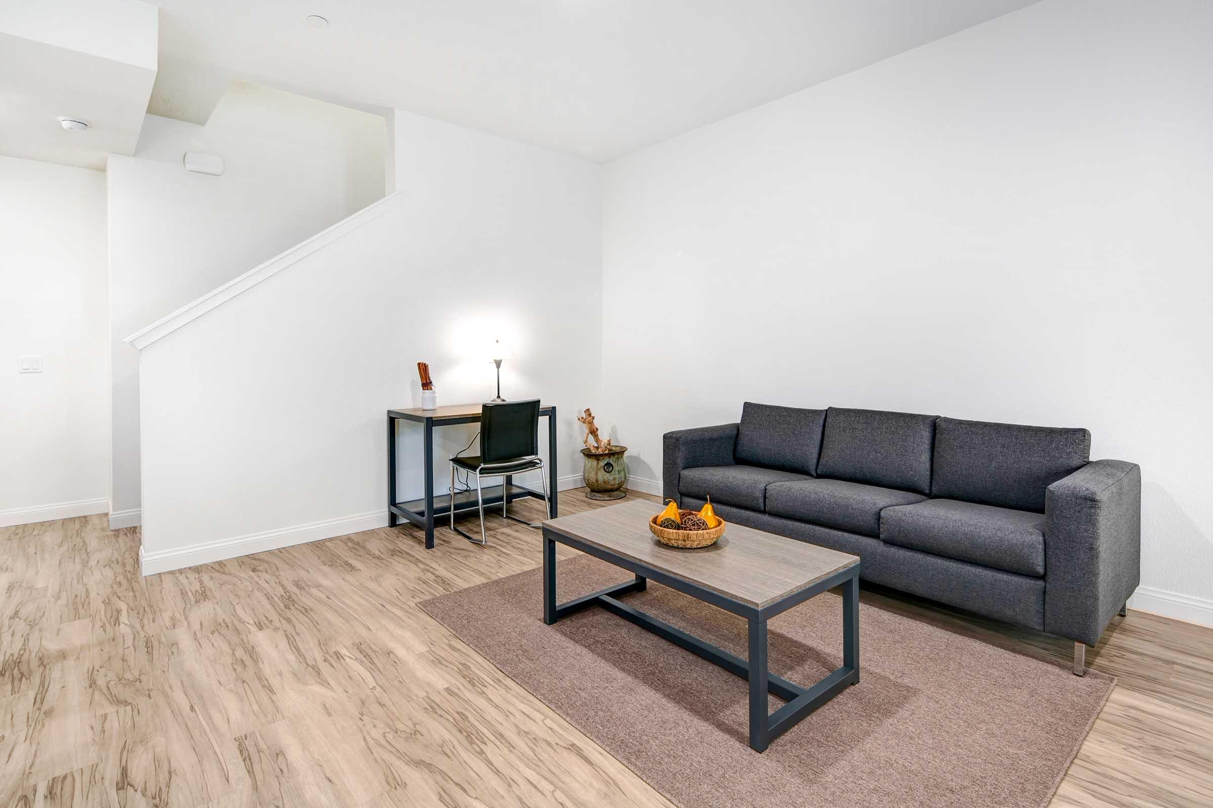 A modern living room featuring a dark gray sofa, a wooden coffee table with decorative items, a small desk with a lamp, and a light-colored rug on a wooden floor. A staircase is visible in the background.