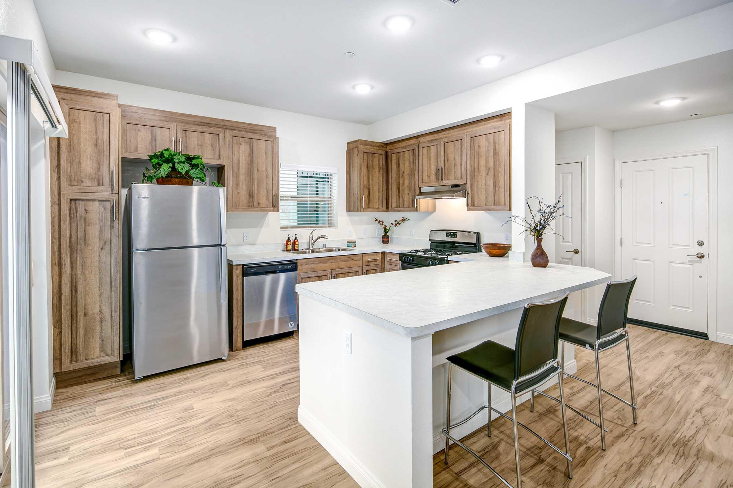 Modern kitchen featuring wooden cabinets, stainless steel appliances including a refrigerator and dishwasher, a countertop with bar seating, and neutral-colored walls and flooring. Natural light enters through a window, enhancing the inviting atmosphere.