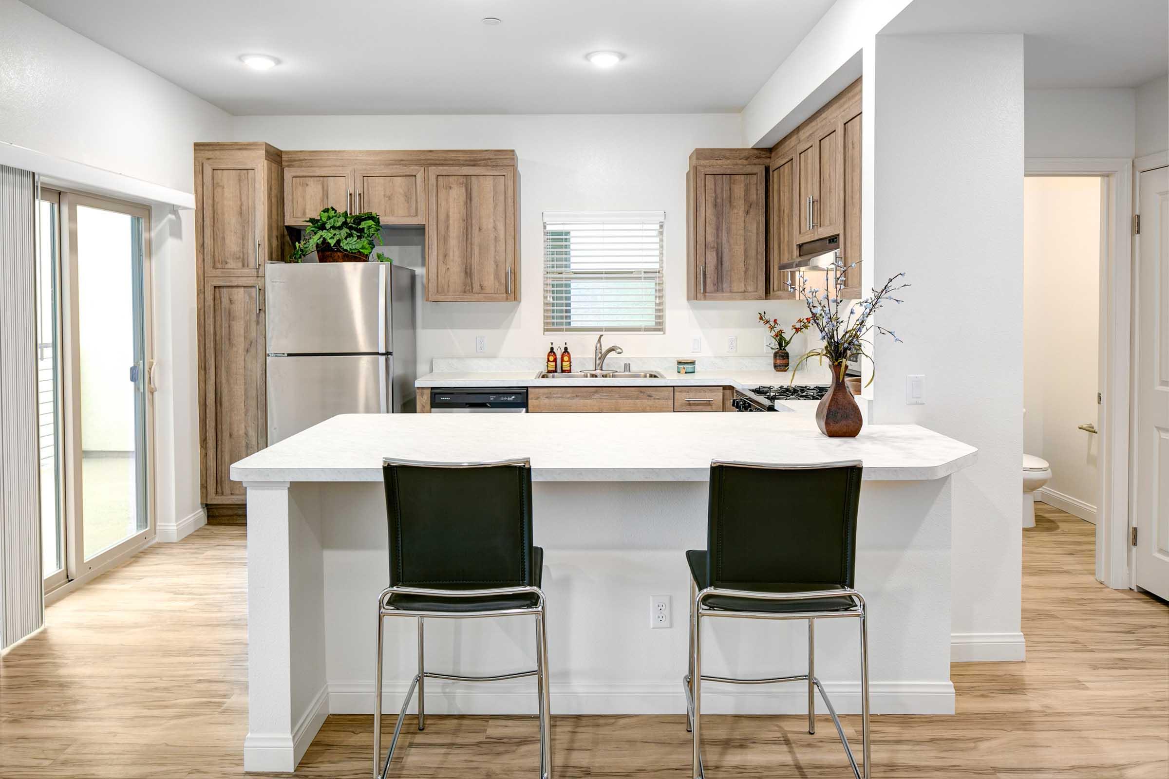 Modern kitchen featuring wooden cabinets, stainless steel appliances, a countertop with bar seating, and natural light coming through a sliding glass door.