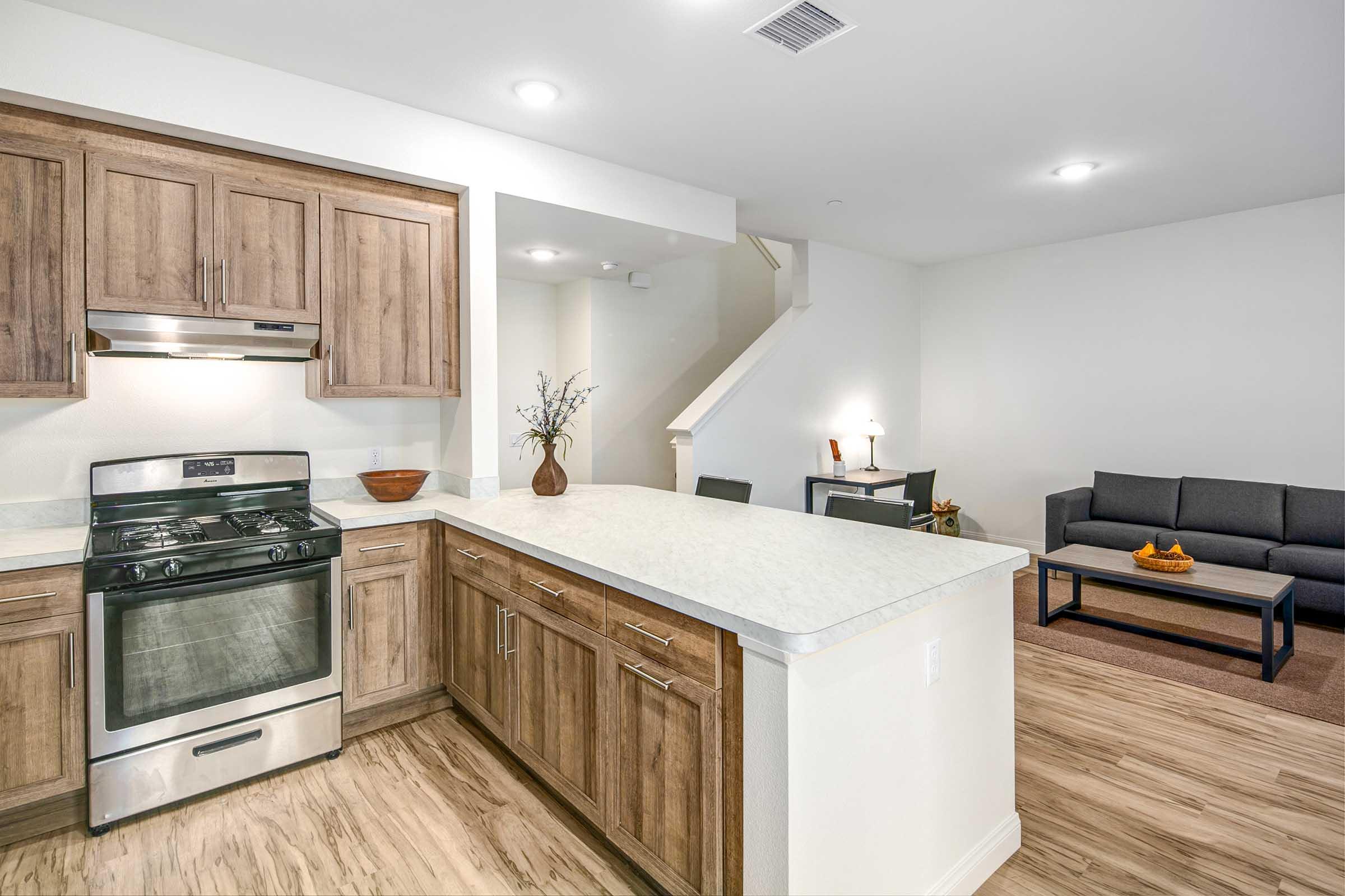 Modern kitchen with wooden cabinetry, a stainless steel gas stove, and a countertop, leading into a light-filled living area featuring a gray sofa, a small coffee table, and a decorative plant. A staircase is visible in the background.