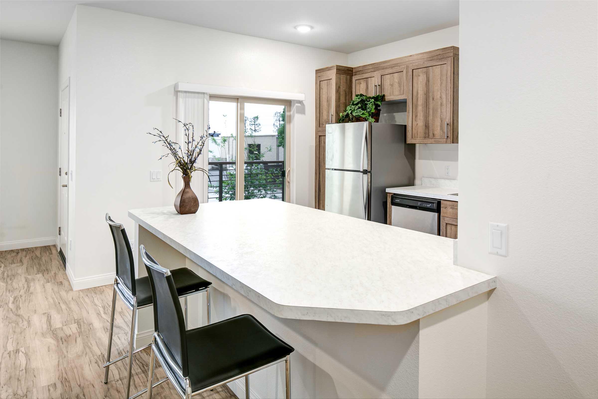 Modern kitchen with a light-colored countertop, dark wood cabinetry, and stainless steel appliances. There are black bar stools at the counter, and a window allows natural light to fill the space. A small vase with branches adds a decorative touch.