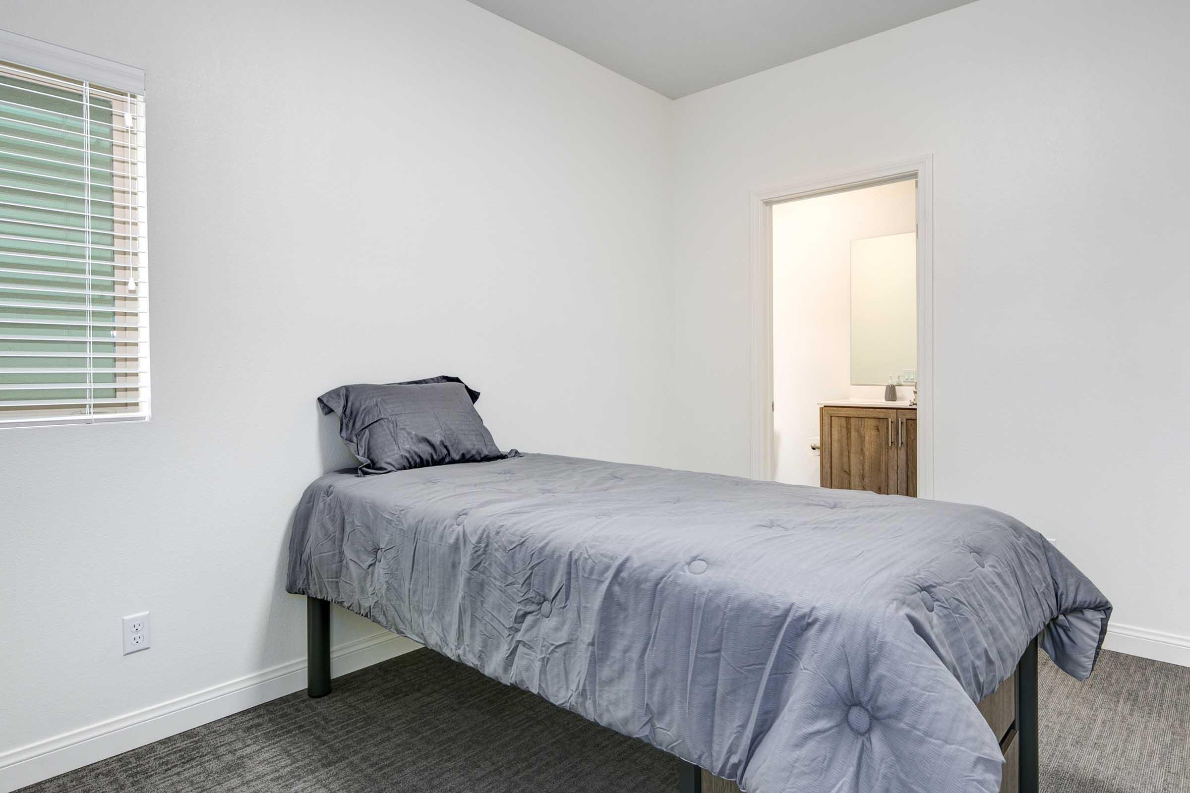 A minimalist bedroom featuring a single bed with gray bedding, positioned against a light-colored wall. A doorway leads to a bathroom, and a window with blinds allows natural light to enter the room.