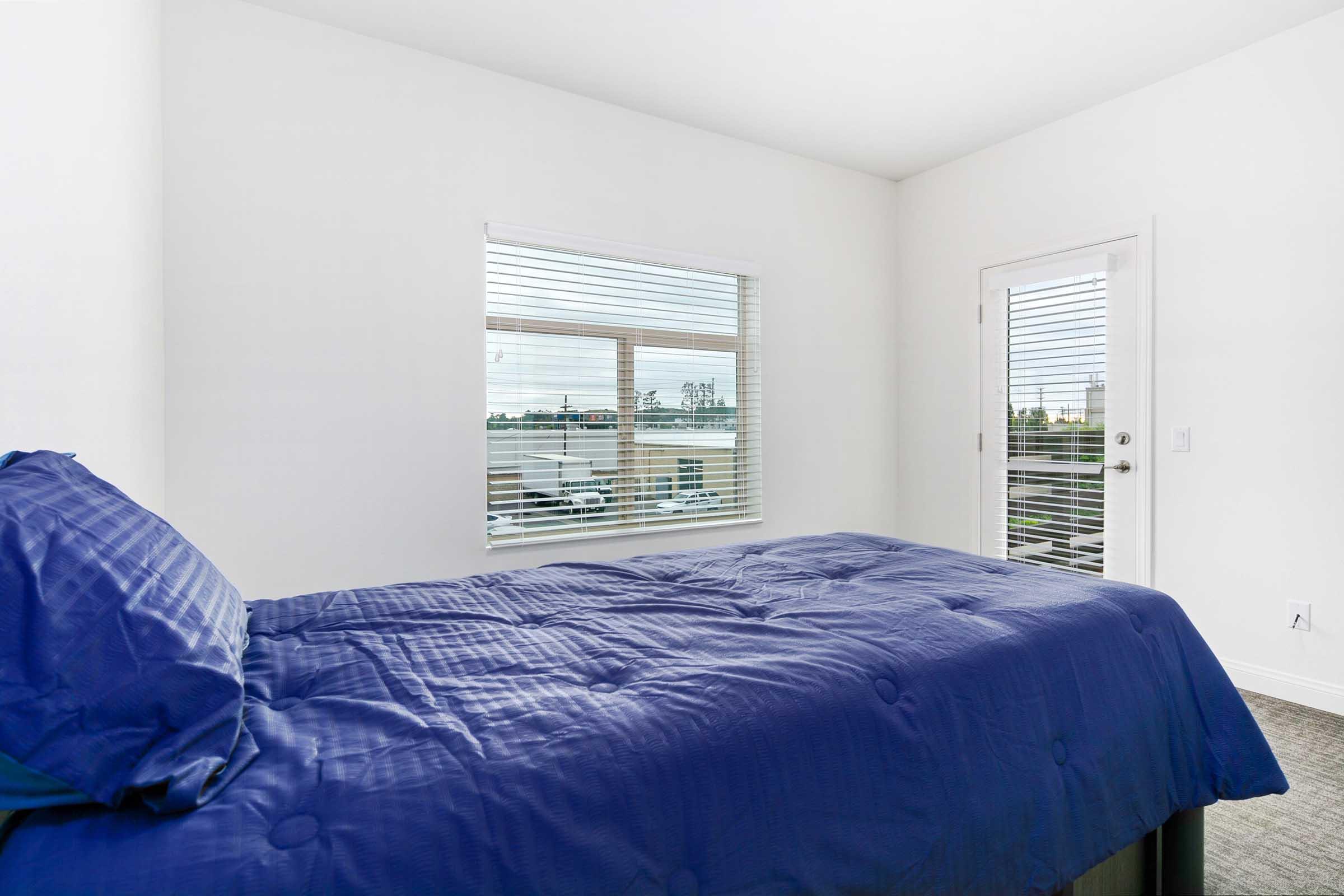 A simple bedroom featuring a bed with a blue comforter, a window with blinds allowing natural light in, and a door leading to a balcony or outdoor area. The walls are painted white, creating a bright and airy atmosphere.