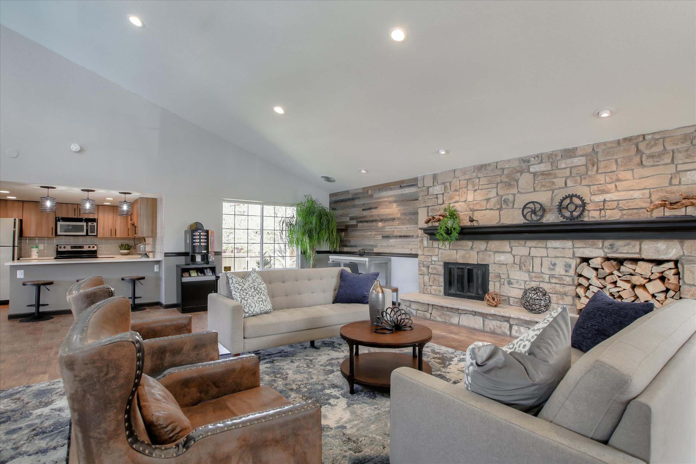 A modern living room featuring a cozy seating area with two gray sofas and brown armchairs, a stone fireplace, and a wooden mantel. Natural light floods the space through large windows, and a stylish coffee table adds a decorative touch. The décor includes plants and soft textiles, creating an inviting atmosphere.