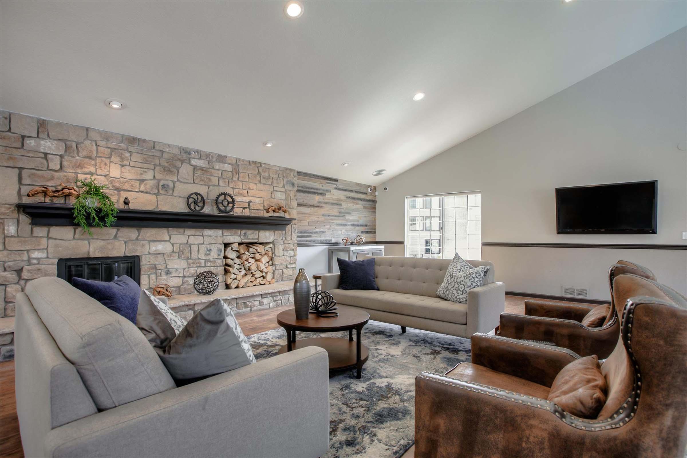 A modern living room featuring a stone fireplace with stacked wood, a plush gray sofa, and two brown leather chairs. A round black coffee table sits in the center on a decorative rug. Natural light streams through a window, illuminating the cozy space, which also includes a mounted television on the wall.