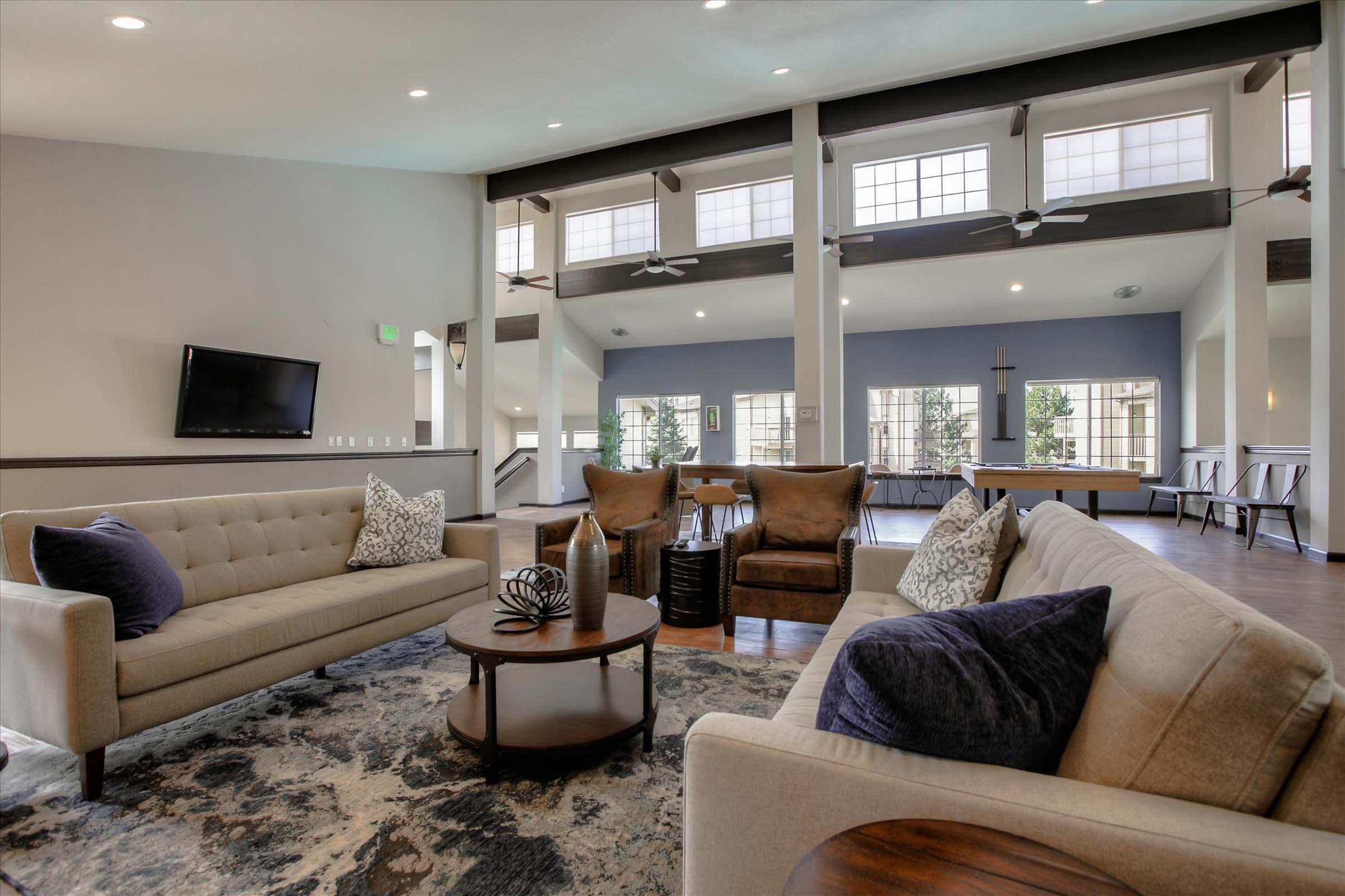 A modern living area featuring a light-colored sectional sofa with decorative pillows, two brown armchairs, and a round coffee table with decorative items. Large windows let in natural light, complementing the neutral color scheme and stylish decor. A flat-screen TV is mounted on the wall, enhancing the contemporary feel.