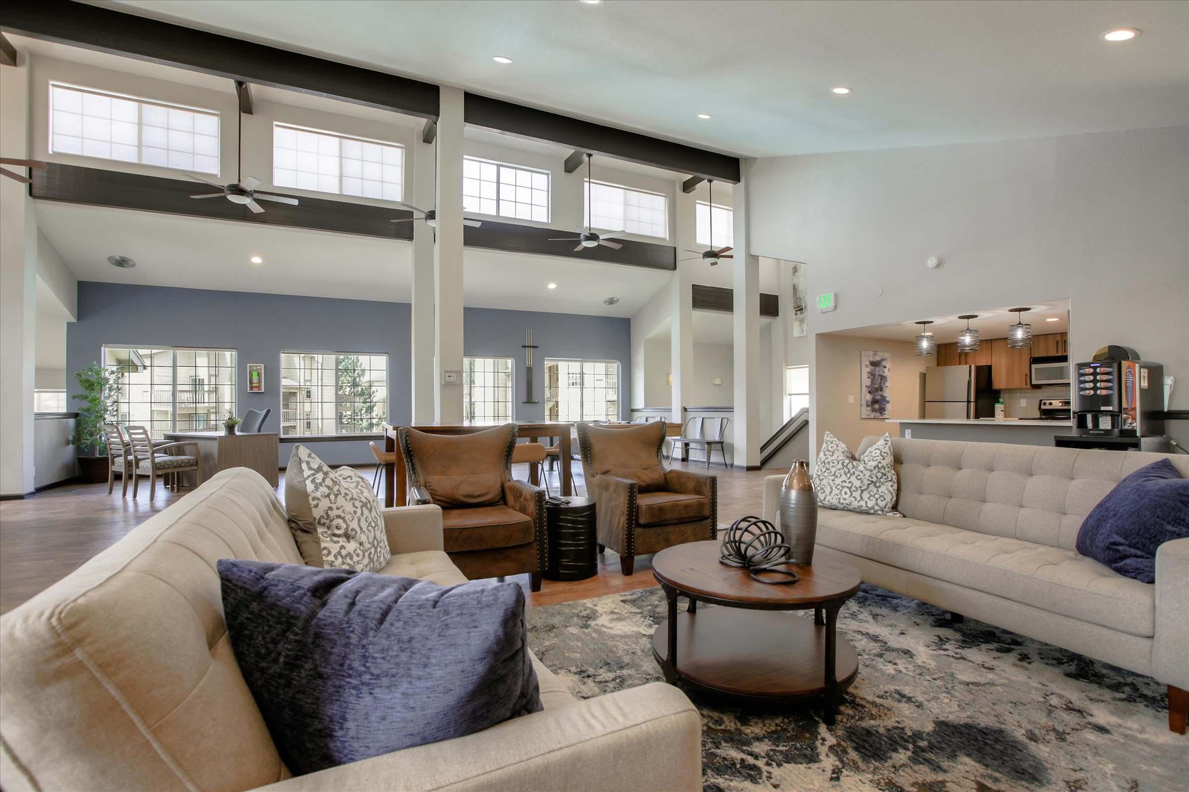 A modern living area featuring a light-colored sectional sofa with decorative pillows, a round coffee table, and a patterned area rug. The space includes large windows allowing natural light, polished wooden floors, and a dining area in the background with contemporary furnishings.