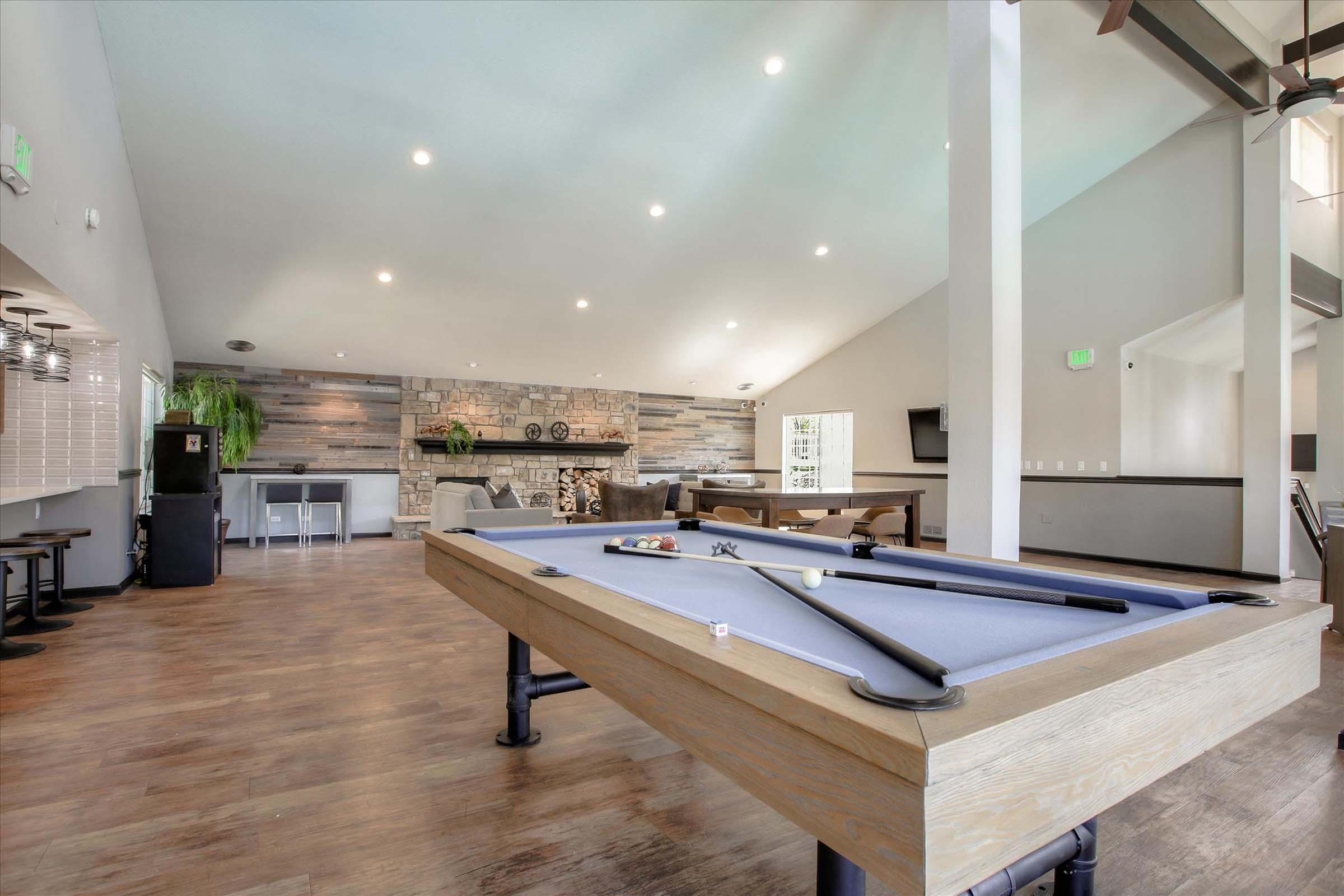 Interior view of a spacious recreational room featuring a pool table in the foreground, a stone fireplace with a modern texture in the background, and ample seating. Bright overhead lighting enhances the open atmosphere, with plants and a large TV visible on the walls.