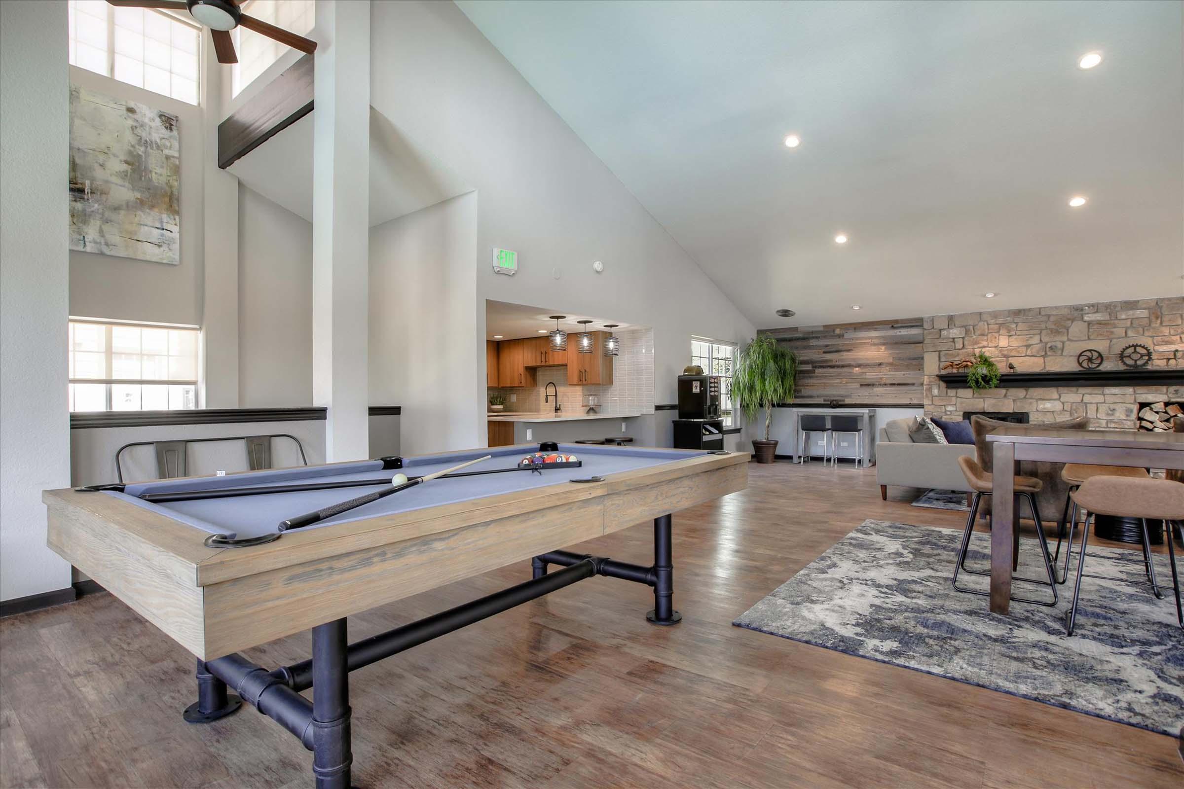 A modern living space featuring a pool table in the foreground, a cozy seating area with a stone fireplace, and a dining table. Large windows allow natural light to fill the room, while stylish decor elements create a welcoming atmosphere.