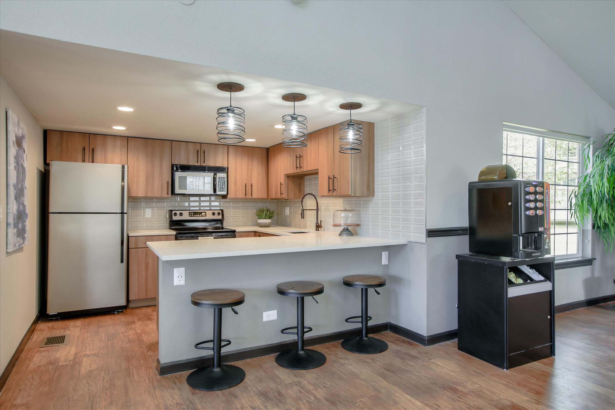 A modern kitchen featuring a stainless steel fridge and stove, wooden cabinetry, a white countertop with bar seating for three, stylish pendant lighting, and a green plant accent. There is also a dark cabinet near a window providing natural light.