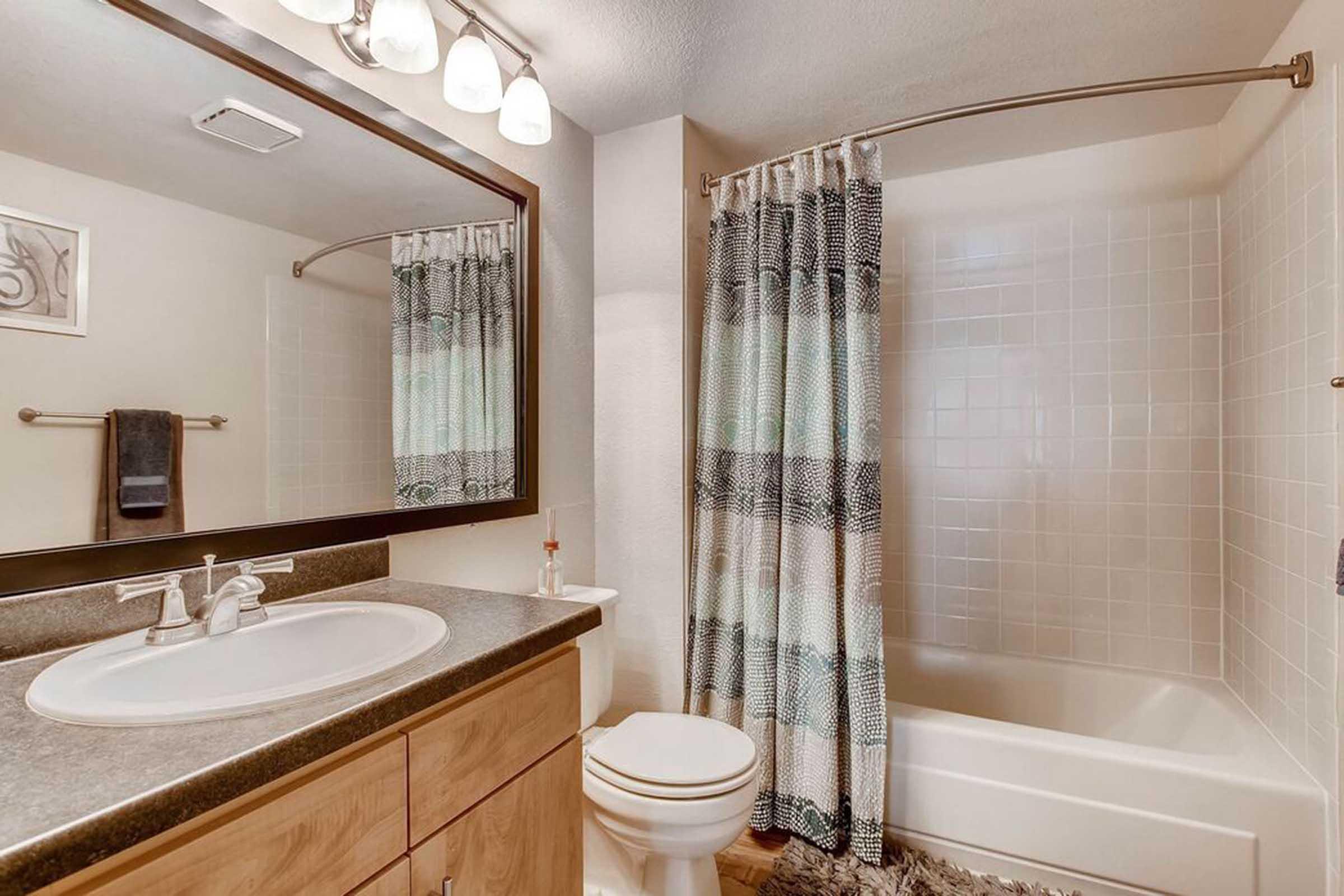 A modern bathroom featuring a freestanding bathtub with a shower, a large mirror above the sink, wood cabinetry, and a patterned shower curtain. Soft lighting from ceiling fixtures enhances the bright, neutral color scheme. A towel rack and a plush rug complete the relaxed ambiance.