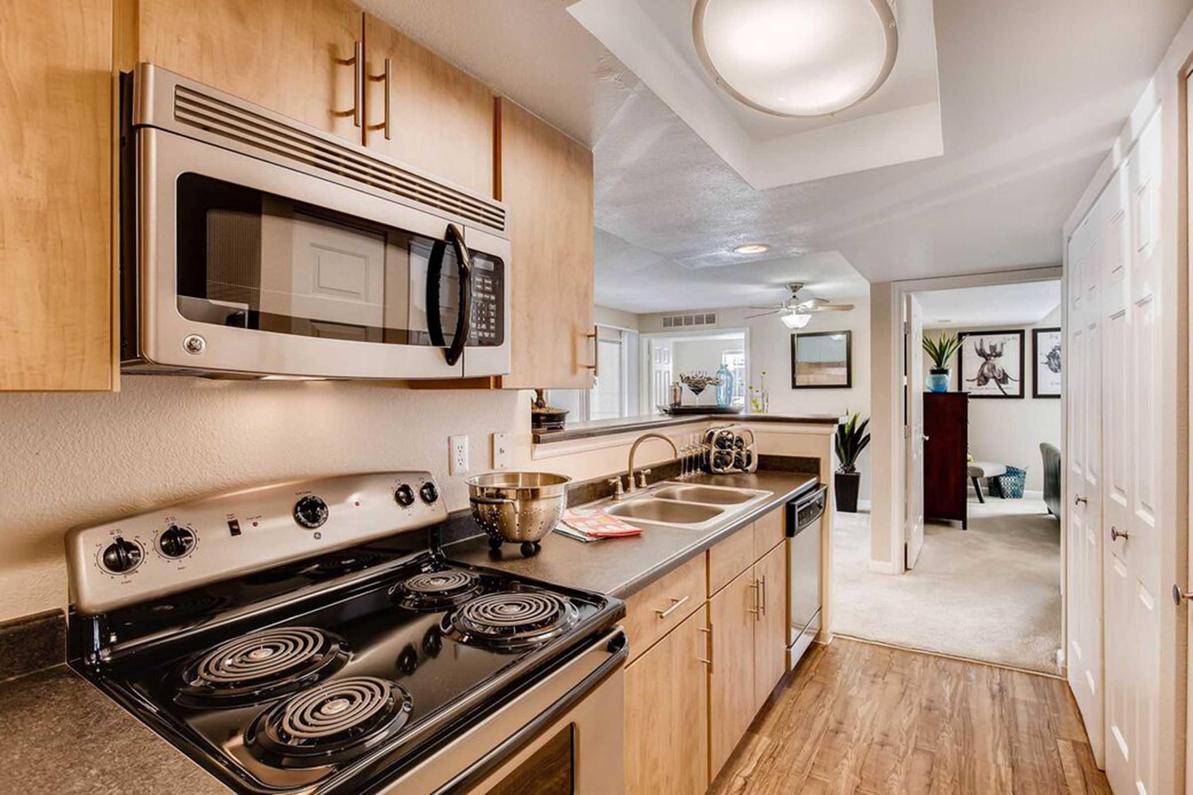 Modern kitchen featuring a microwave above a stove, a dual sink, and wooden cabinetry. The countertop is dark, with a stainless steel bowl on it. The background shows an open layout leading to a living area with decor visible. Bright lighting enhances the space's inviting atmosphere.