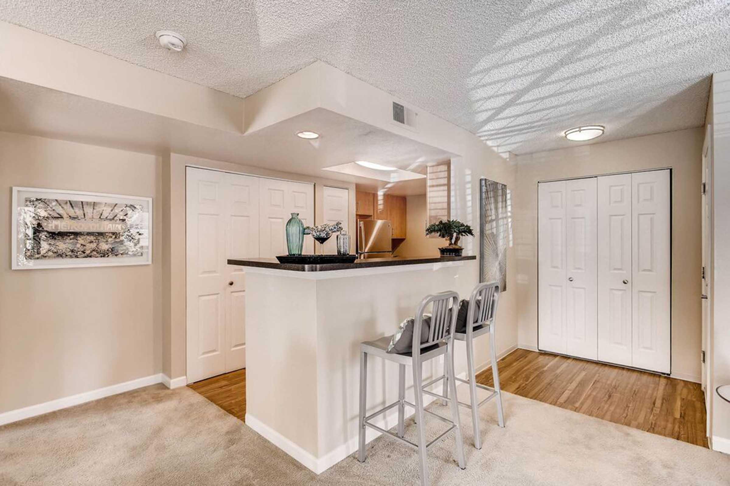 A modern interior featuring a cozy entryway with a small bar area and two stools. Light-colored walls complement the beige carpet. There are two white doors leading to closets. Decorative items on the bar add a touch of style, and soft lighting creates a warm atmosphere.