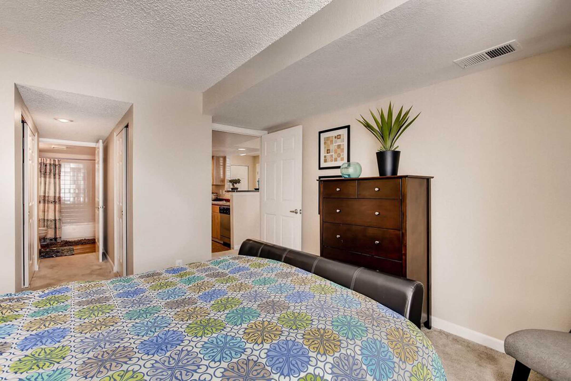 A cozy bedroom featuring a neatly made bed with a colorful patterned quilt. A tall dresser stands against the wall, accompanied by a potted plant on top. The room has soft lighting and adjacent doorways lead to other spaces, with a glimpse of a kitchen area in the background.
