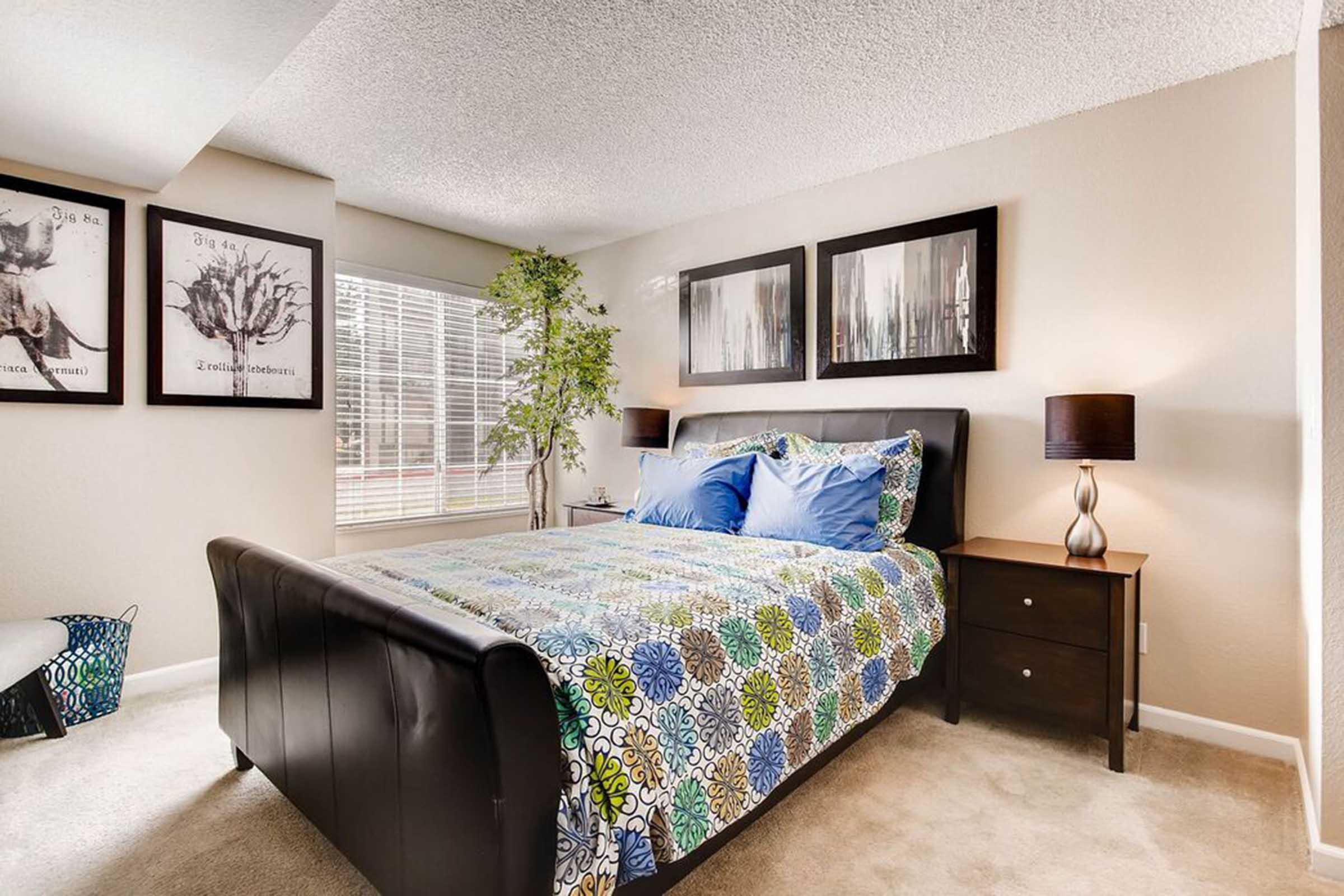 A cozy bedroom featuring a queen-sized bed with a colorful patterned quilt, decorative pillows, and stylish black furniture. There are two framed artworks on the wall, a potted plant, and a bedside table with a lamp. Large windows allow natural light to brighten the space.