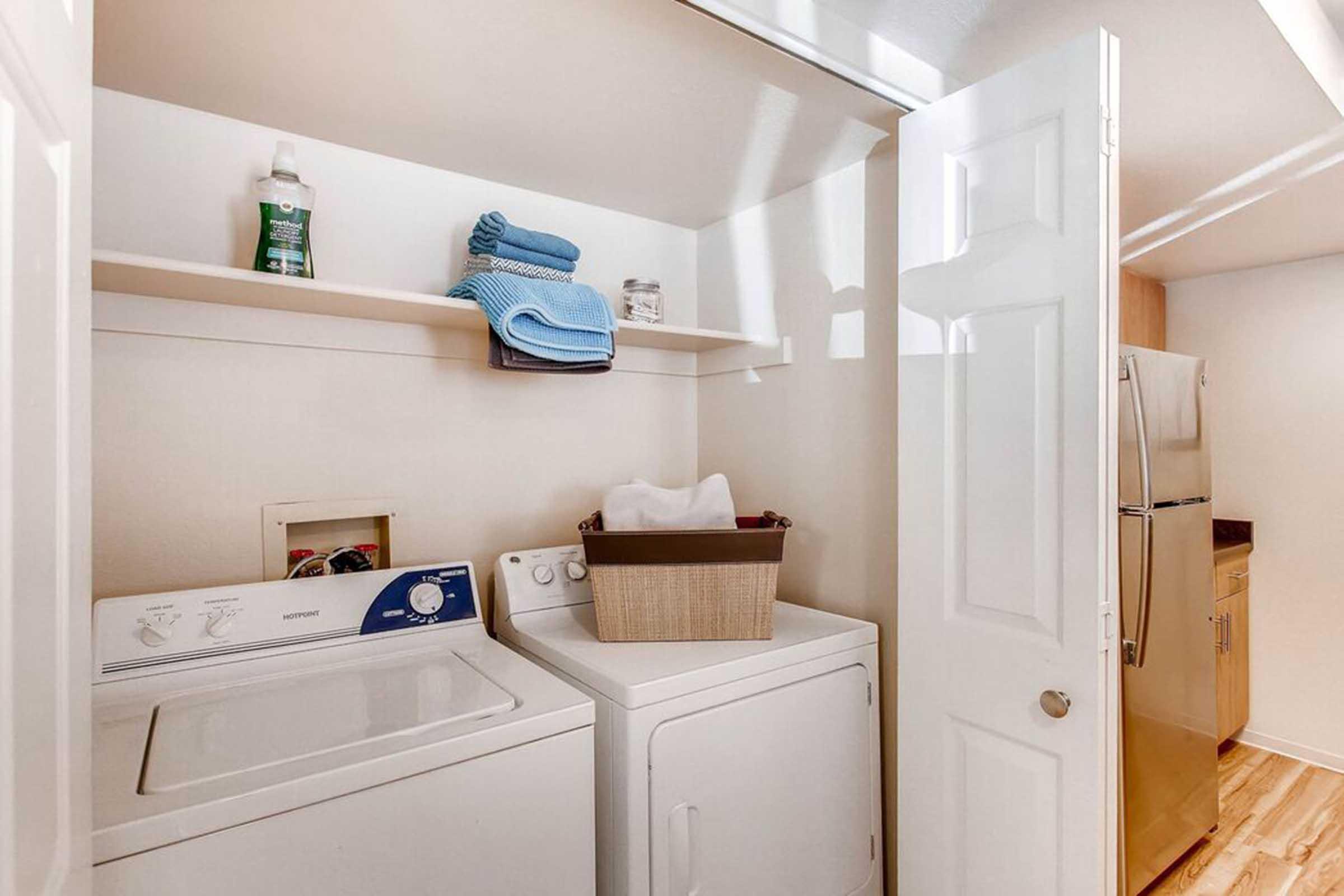 A laundry room with a washing machine and dryer side by side. Above, there are shelves holding cleaning supplies and neatly folded towels. A basket sits on the dryer, and a partially open door leads to another area, with a glimpse of a refrigerator at the right. The floor is wood laminate.