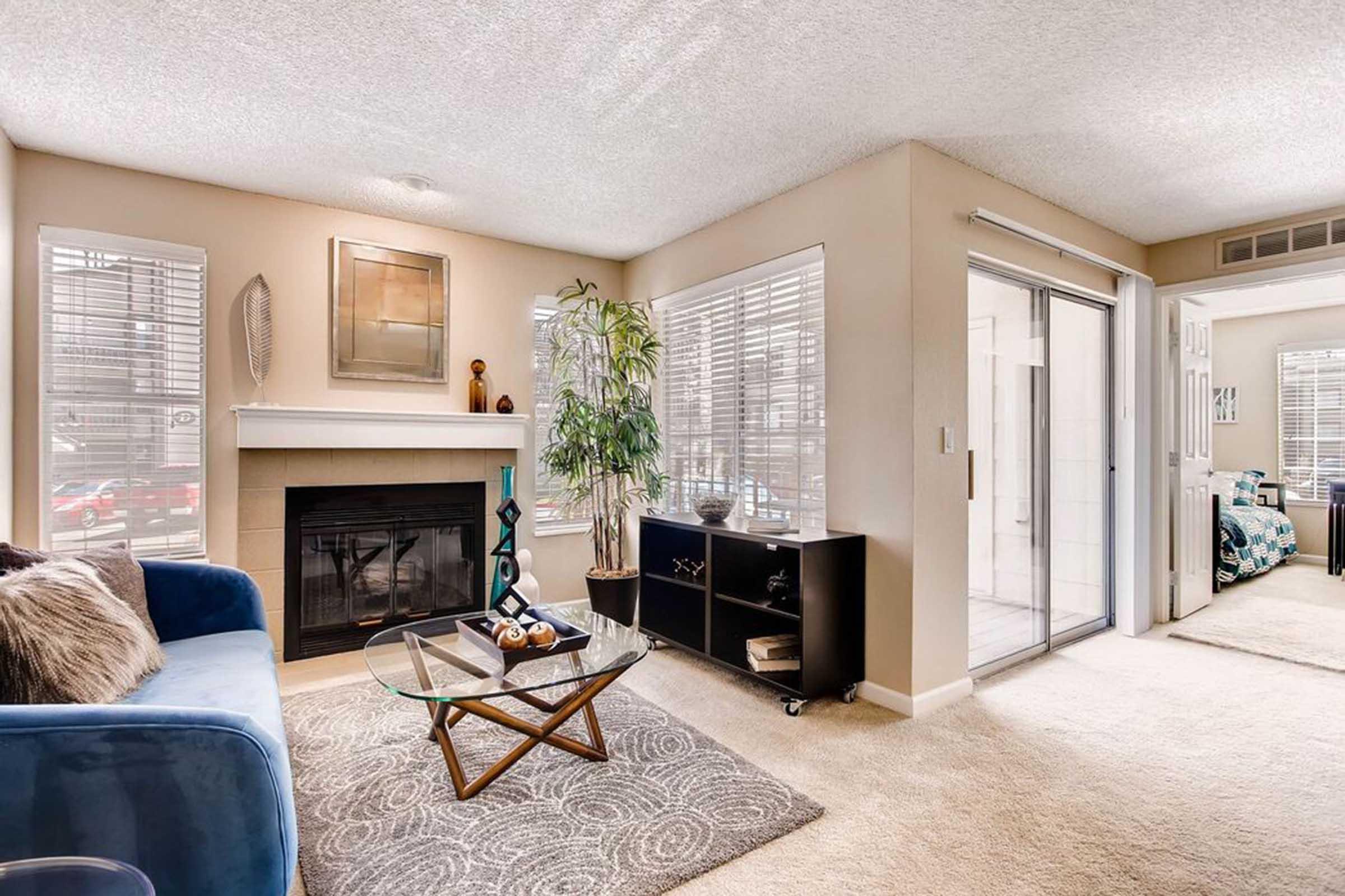 Cozy living room featuring a blue sofa, a glass coffee table, and a decorative fireplace. Large windows allow natural light, and there’s a plant near the corner. A black cabinet displays decorative items. The room leads to a sliding glass door with a view of a bright, airy space beyond.