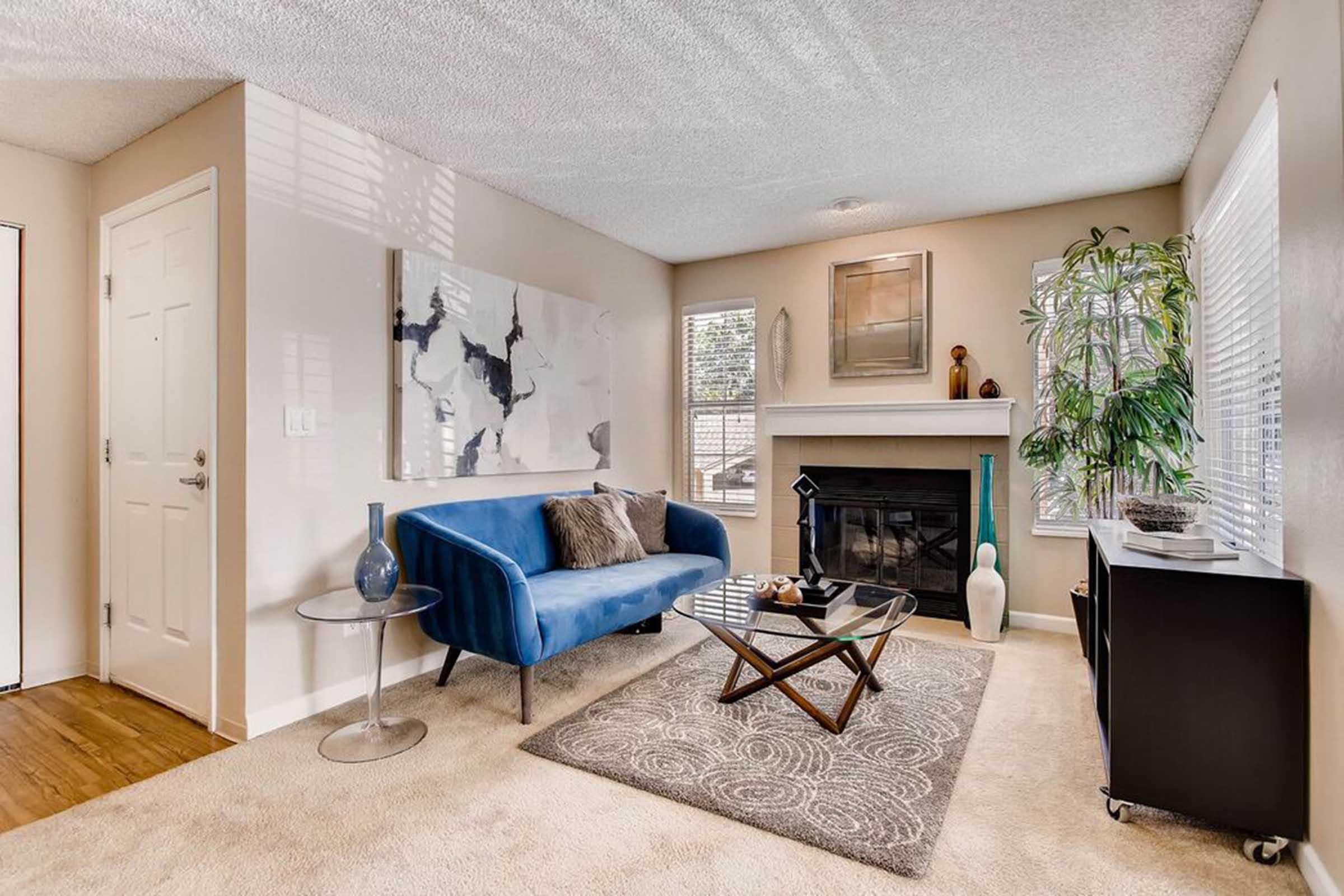 A modern living room featuring a blue velvet sofa, a glass coffee table, and a fireplace. The walls are painted in neutral tones, with a large abstract artwork above the fireplace. Large windows let in natural light, and a tall green plant adds a touch of color. Soft carpet covers the floor, creating a cozy atmosphere.