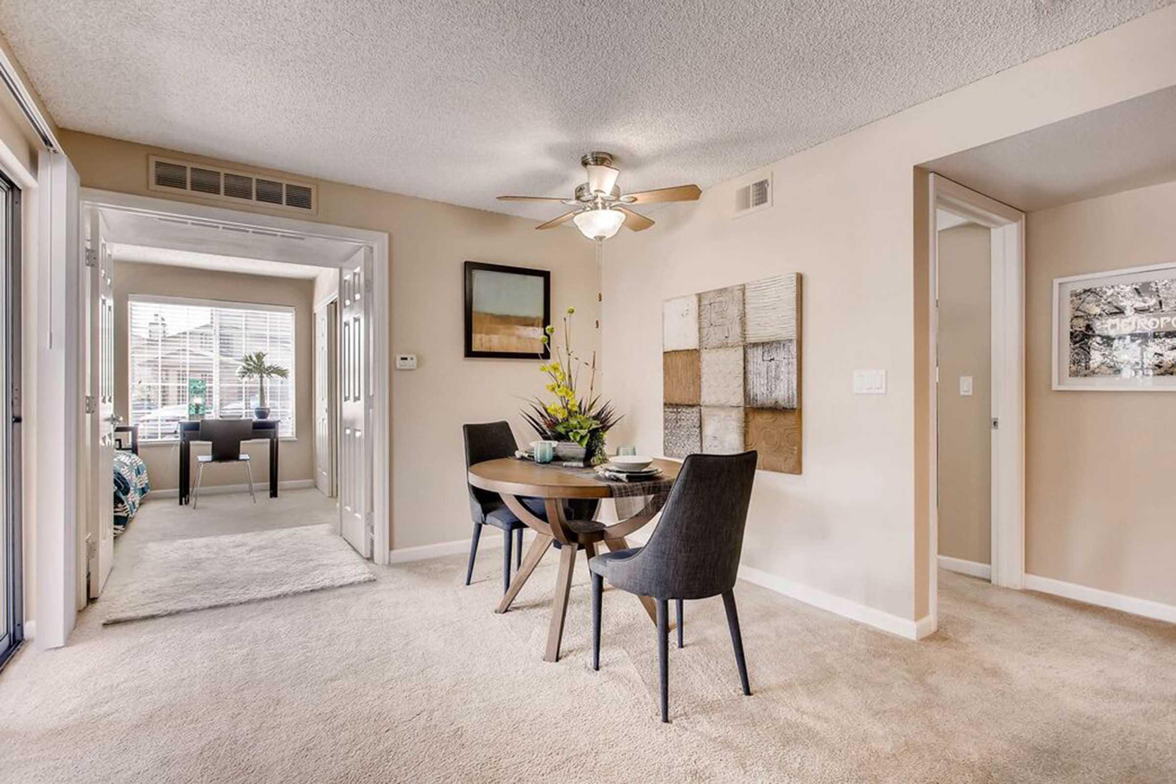 A cozy dining area featuring a round wooden table with four chairs. The room has beige walls, a ceiling fan, and a decorative arrangement of flowers on the table. A doorway leads to a well-lit living space with large windows, and an art piece is displayed on the wall nearby. The flooring is carpeted.
