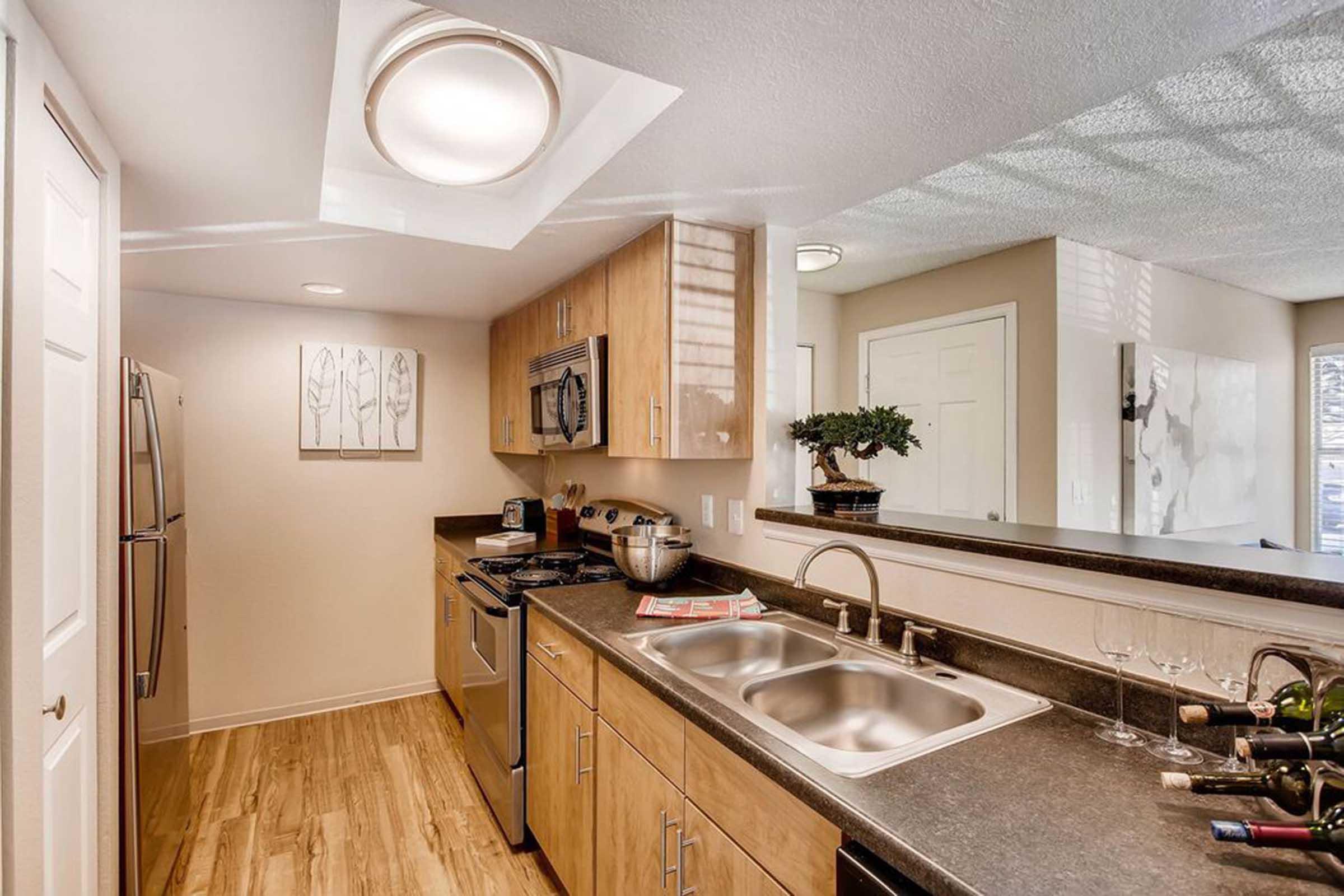 Modern kitchen with wooden cabinets, stainless steel appliances, and a double sink. A bowl and kitchen utensils are on the countertop. There's a small dining area visible in the background, and natural light streams in from a ceiling window.