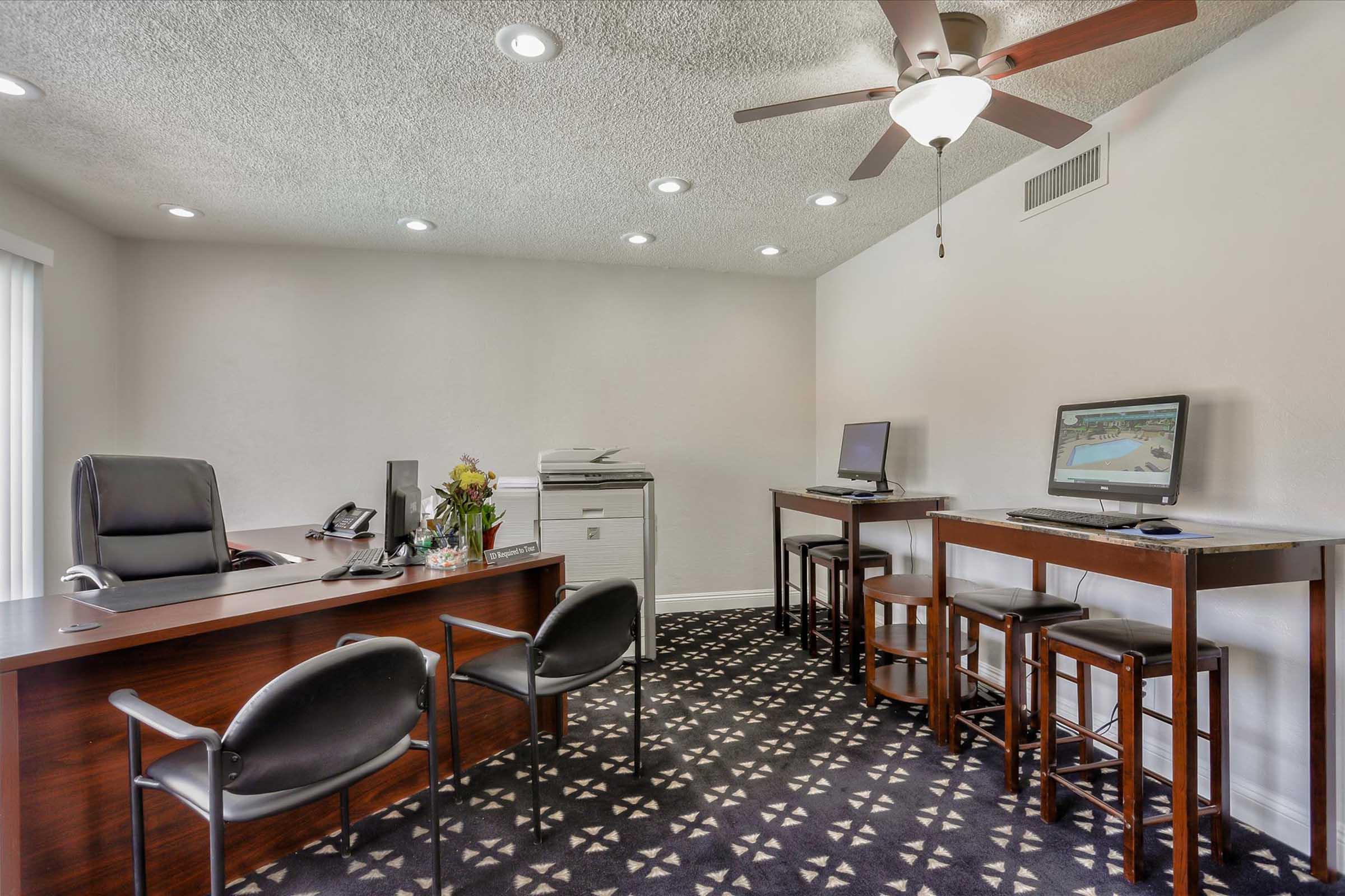 A modern office space featuring a wooden desk with a chair, a printer, and two computers on high tables. There are two additional chairs and bright ceiling lights. A decorative flower arrangement adds a touch of color to the room, which has a patterned carpet and neutral wall colors.