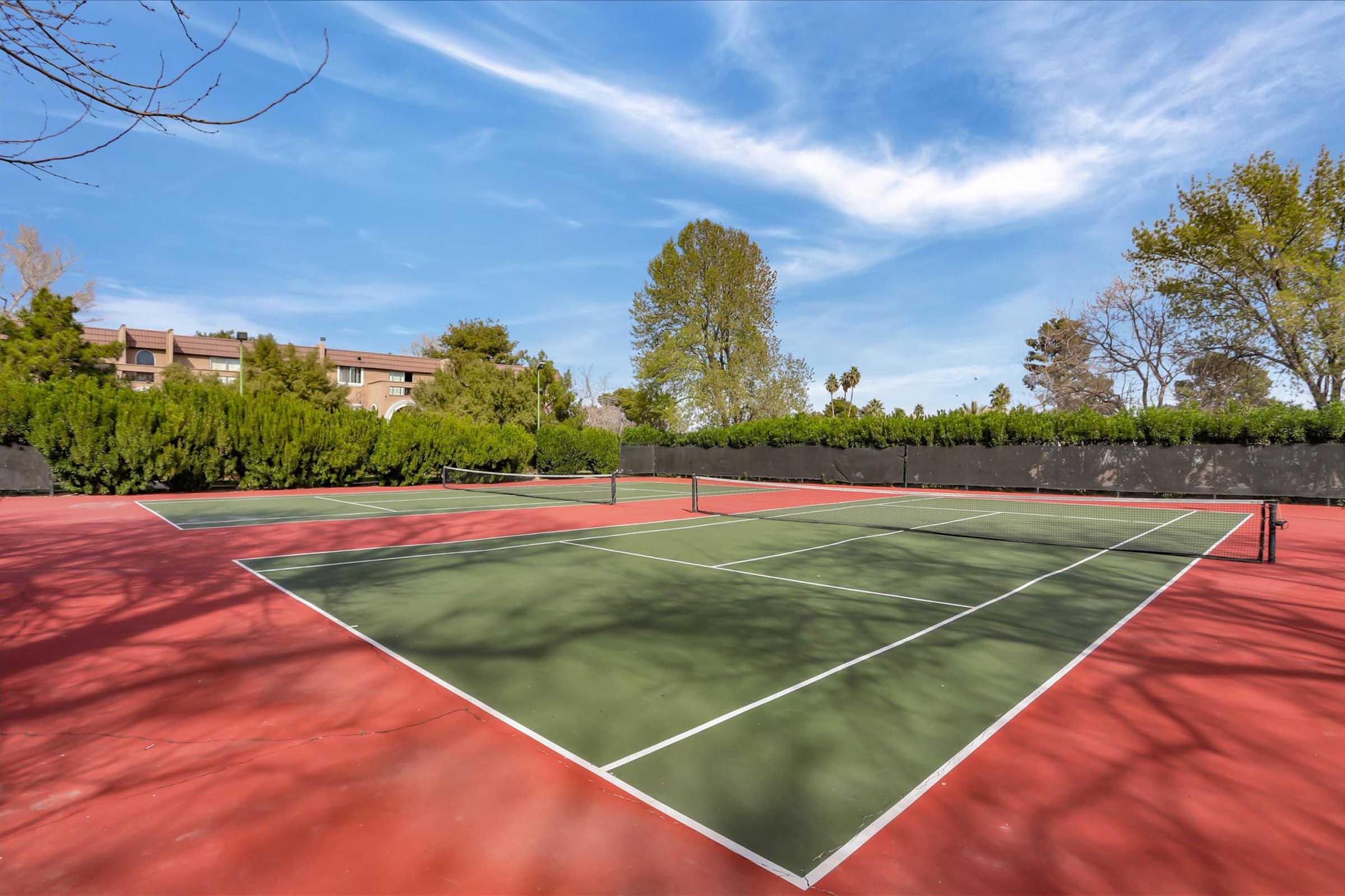 An empty tennis court with a red and green surface, surrounded by greenery and trees. The sky is clear with a few clouds. In the background, there are residential buildings visible behind the hedges. The scene conveys a calm and inviting atmosphere for outdoor sports.