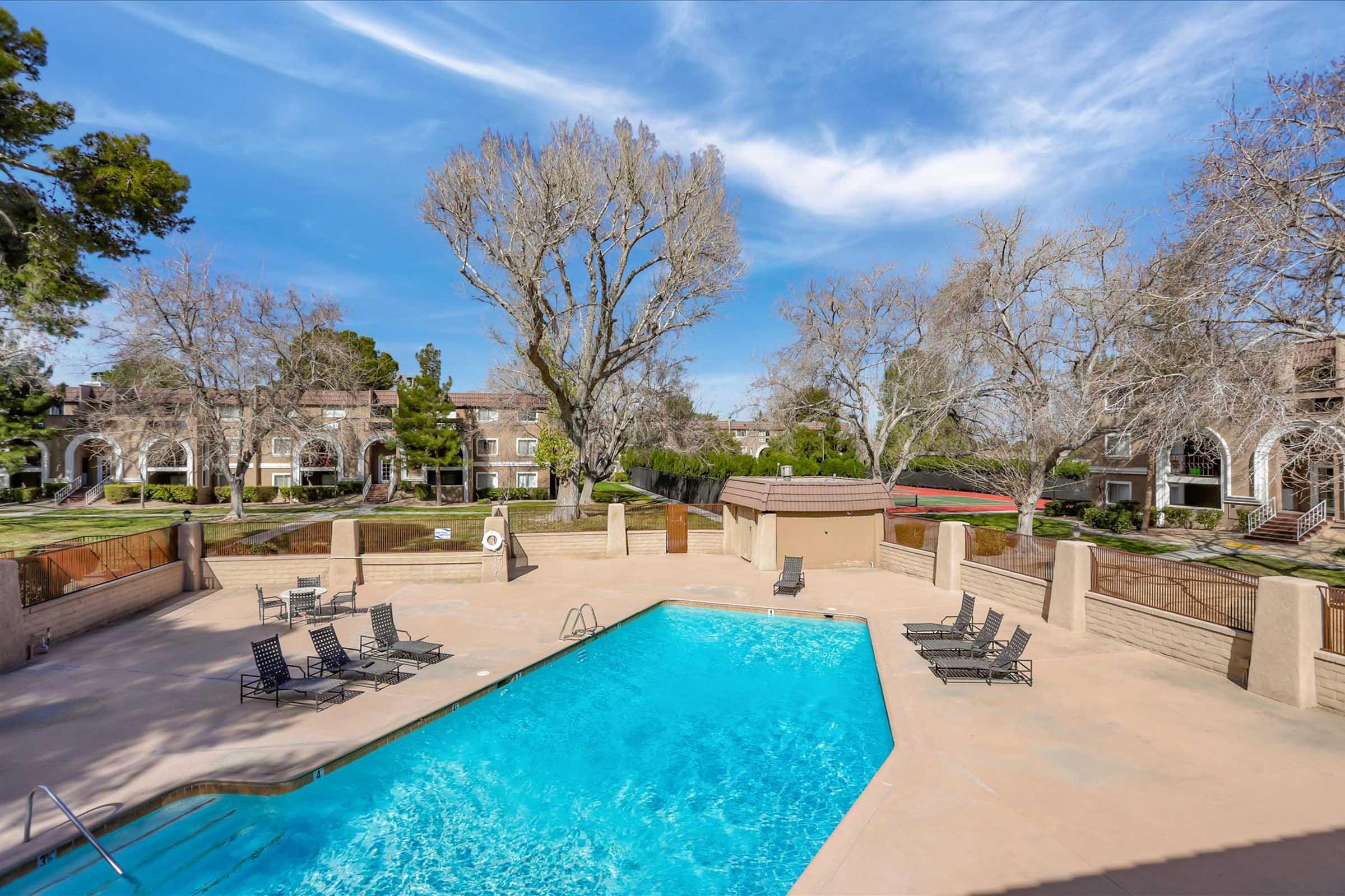 A clear swimming pool surrounded by lounge chairs is set in a picturesque outdoor area with leafless trees and well-maintained landscaping. Apartment buildings are visible in the background under a blue sky with wispy clouds, creating a serene and inviting atmosphere.