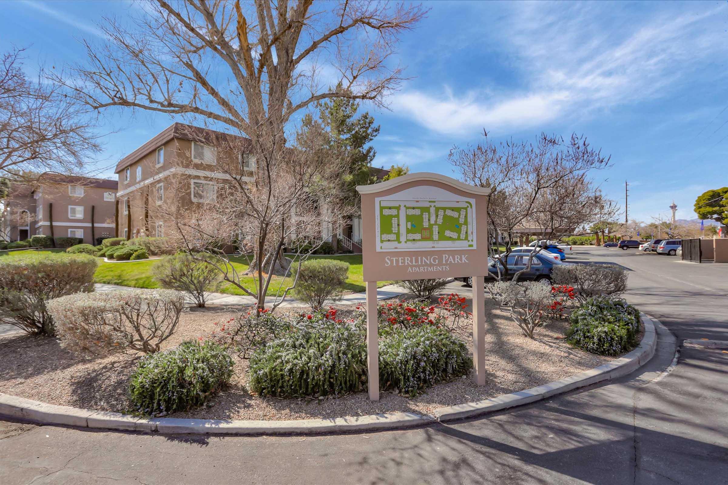 A sign for Sterling Park Apartments is prominently displayed, surrounded by landscaped gardens with shrubs and flowering plants. In the background, there are residential buildings and clear blue skies. The area appears well-maintained and inviting.