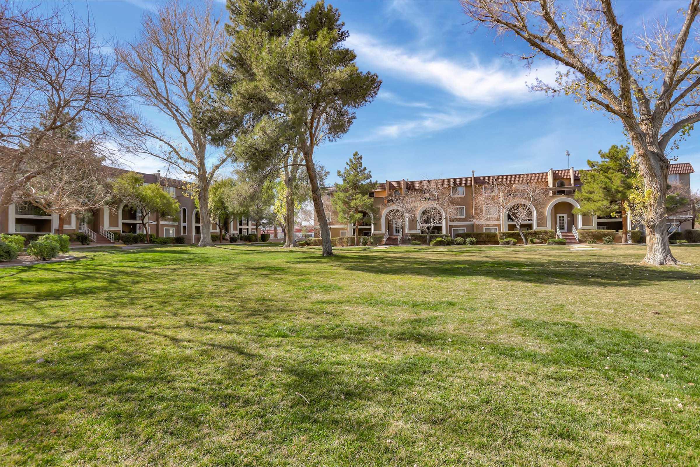 A green lawn surrounded by trees and shrubs, with two-story apartment buildings in the background under a clear blue sky. The scene is bright and open, showcasing well-maintained landscaping.