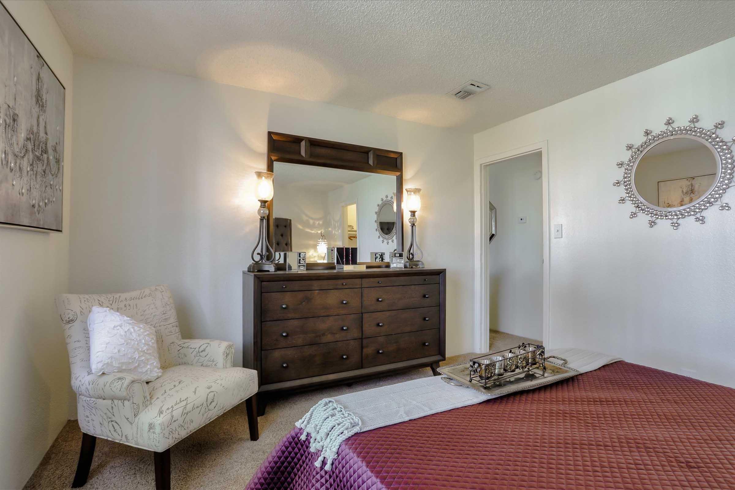 Cozy bedroom featuring a bed with a burgundy quilt, a stylish dresser with decorative lamps, a comfortable chair, and a round mirror on the wall. Soft lighting creates a warm ambiance, enhancing the inviting atmosphere of the room.