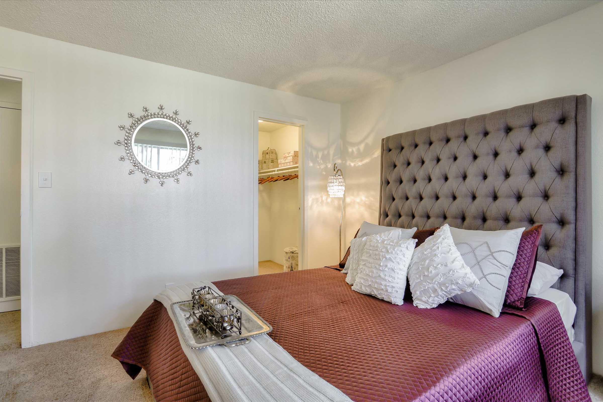 A cozy bedroom featuring a tufted gray headboard, luxurious bedding with decorative pillows, and a stylish round mirror on the wall. A closet is visible in the background, and the room is softly lit, creating an inviting atmosphere. A decorative tray rests on the bed.